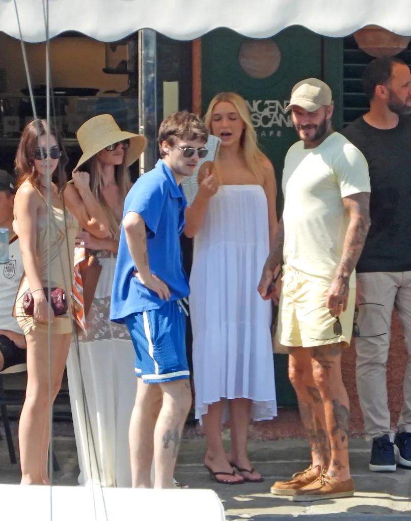 Victoria Beckham wearing a straw hat and sunglasses, Cruz Beckham in a blue outfit, Jackie Apostel in a white dress, and David Beckham in a light yellow t-shirt, shorts, and a beige cap enjoying a family day out.