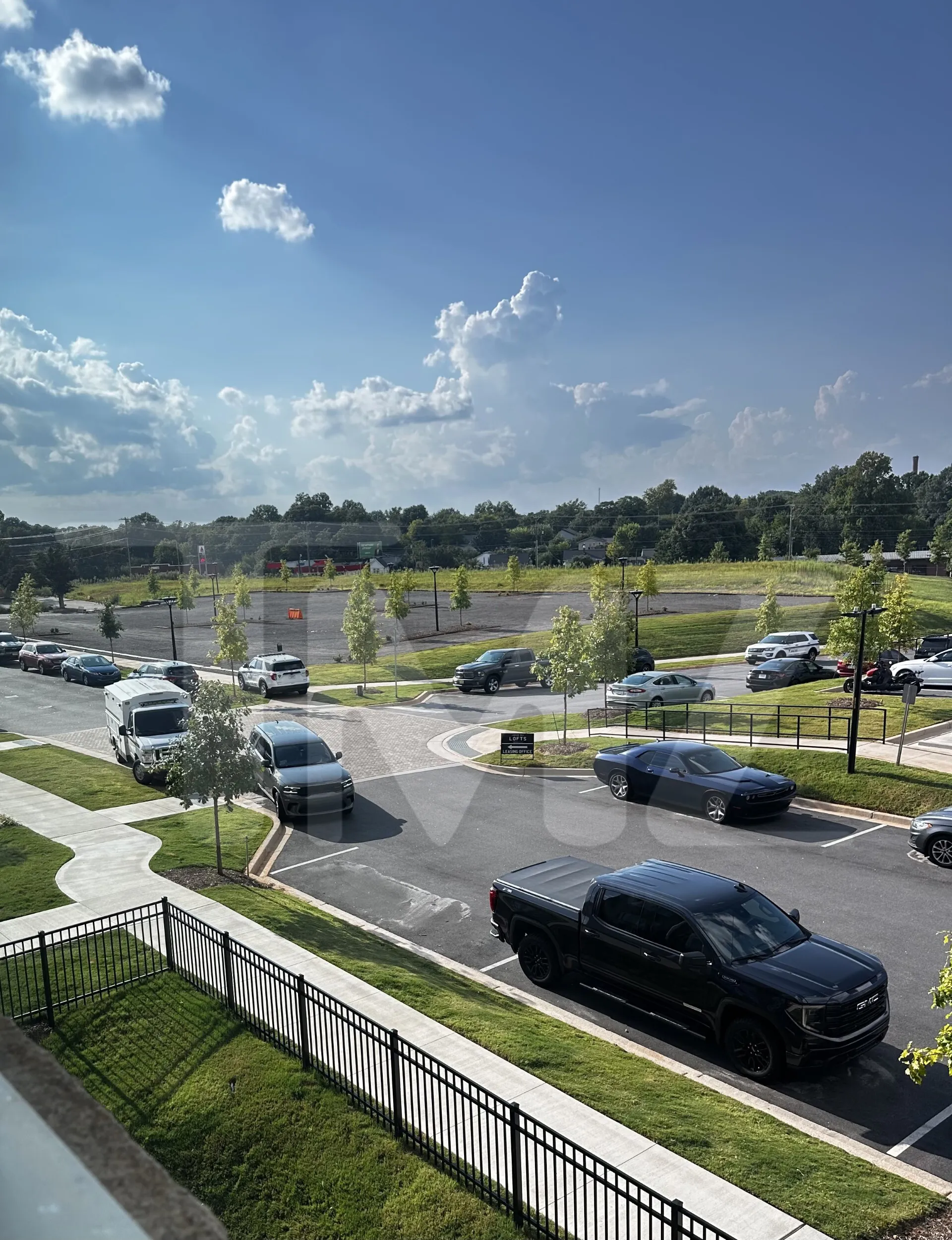 An aerial view of a parking lot with several vehicles, including a white van, a dark grey SUV, and a black pickup truck, under a partly cloudy sky.