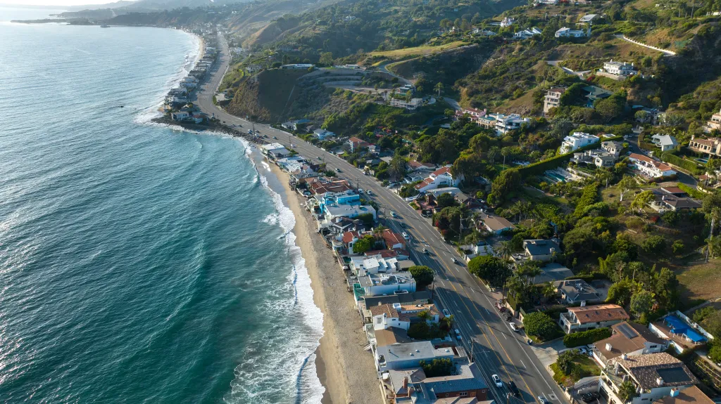 Aerial view of the Pacific Coast Highway in Malibu, California, with the ocean and beach on the left and houses on hills to the right.