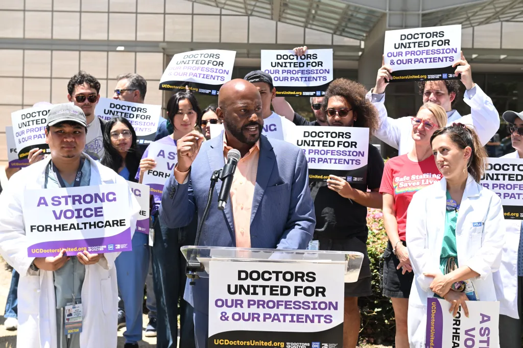 A man speaks at a podium to a crowd of doctors holding signs that read 
