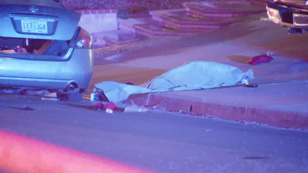 A silver Nissan Altima with its trunk open, and a body on the ground covered by a white sheet, with a red blanket nearby.