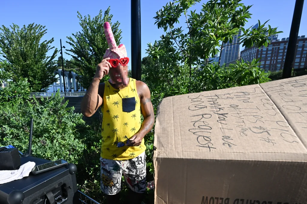 A man wearing a penis-shaped mask and heart-shaped glasses, standing next to a cardboard box with 