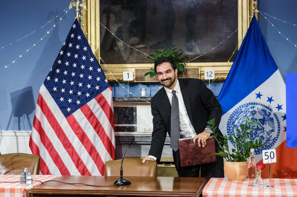 A smiling man in a suit stands between the American flag and the New York City flag.