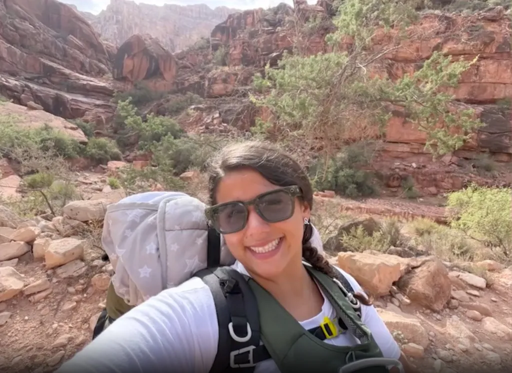 A woman wearing sunglasses, a white shirt, and a baby carrier backpack smiles at the camera on a hiking trail.