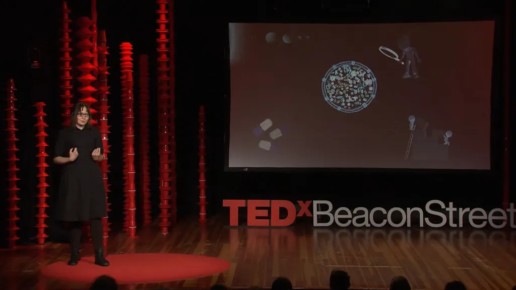 Kate Adamala speaking at a TEDxBeaconStreet event, with a screen displaying illustrations of a synthetic cell, pills, and small figures.