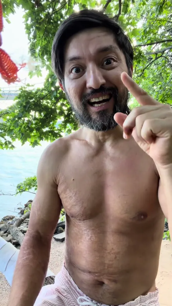 Galymzhan Abaildayev, 44, with a beard and no shirt, standing outdoors next to the water and trees, gesturing with his finger, and looking directly at the camera.