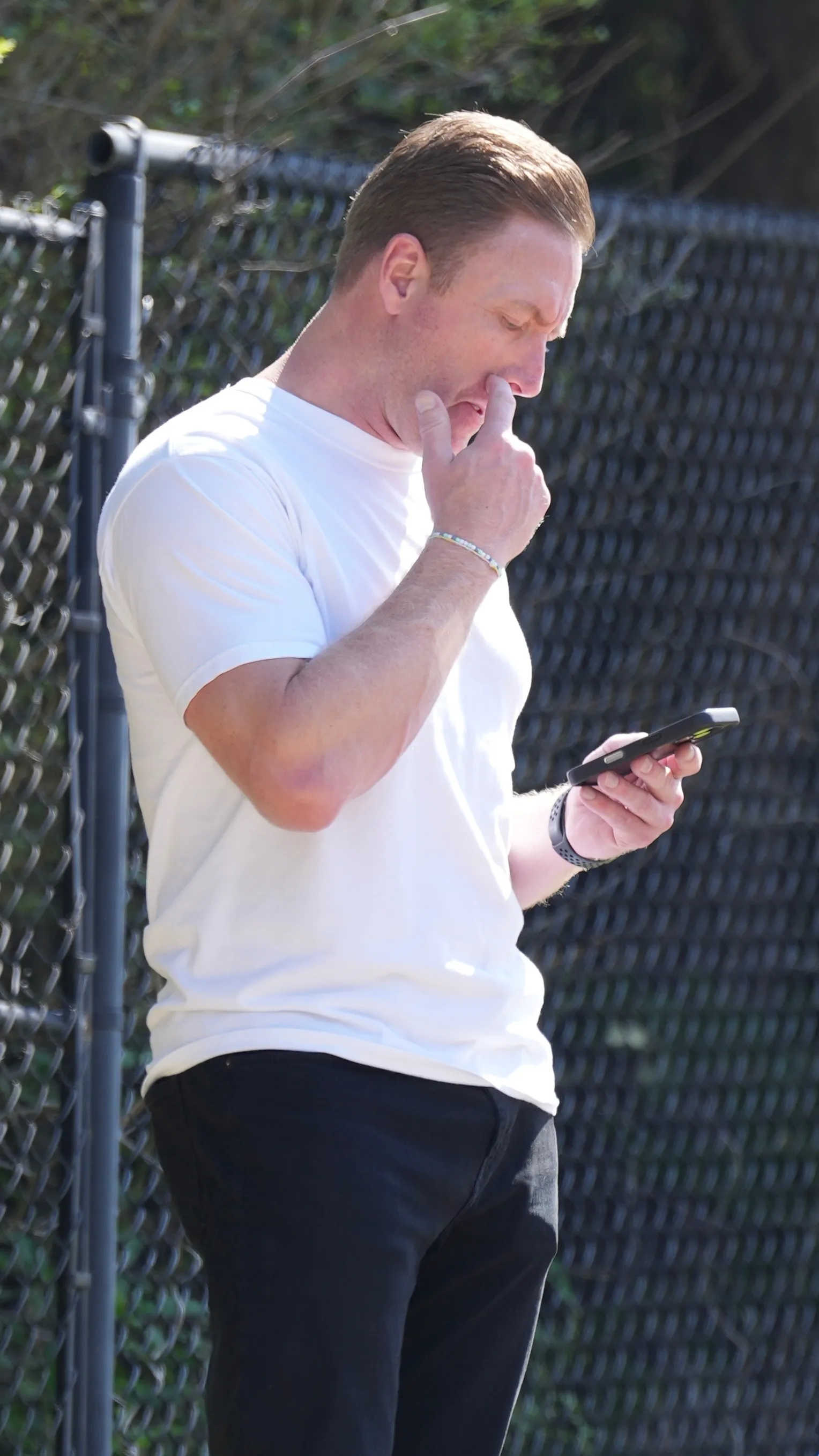 Kroy Biermann in a white t-shirt and dark pants, holding a phone, with one finger near his nose.