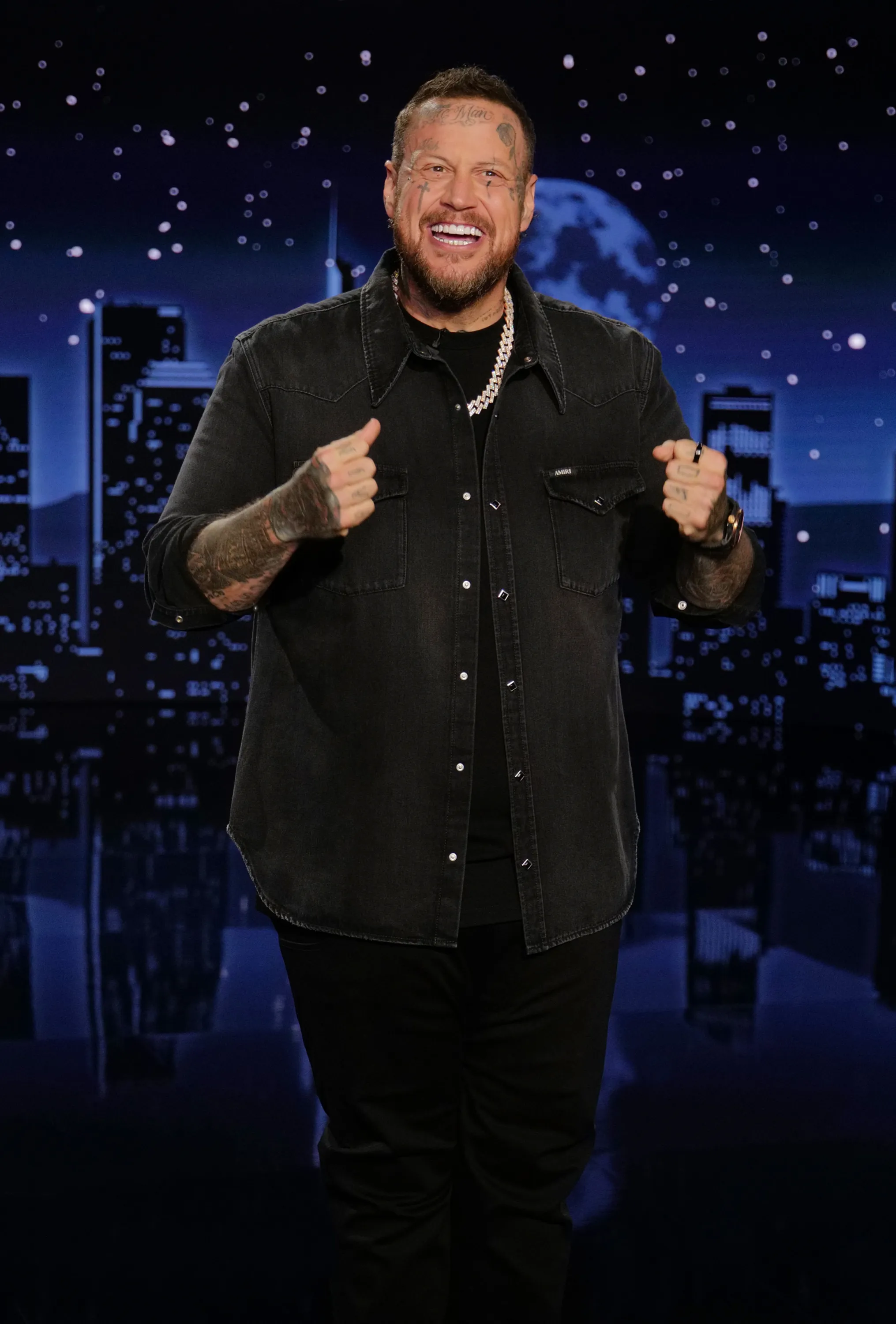 Jelly Roll smiling and pumping his fists, wearing a dark denim shirt and black pants, against a backdrop of a city skyline at night with a full moon.
