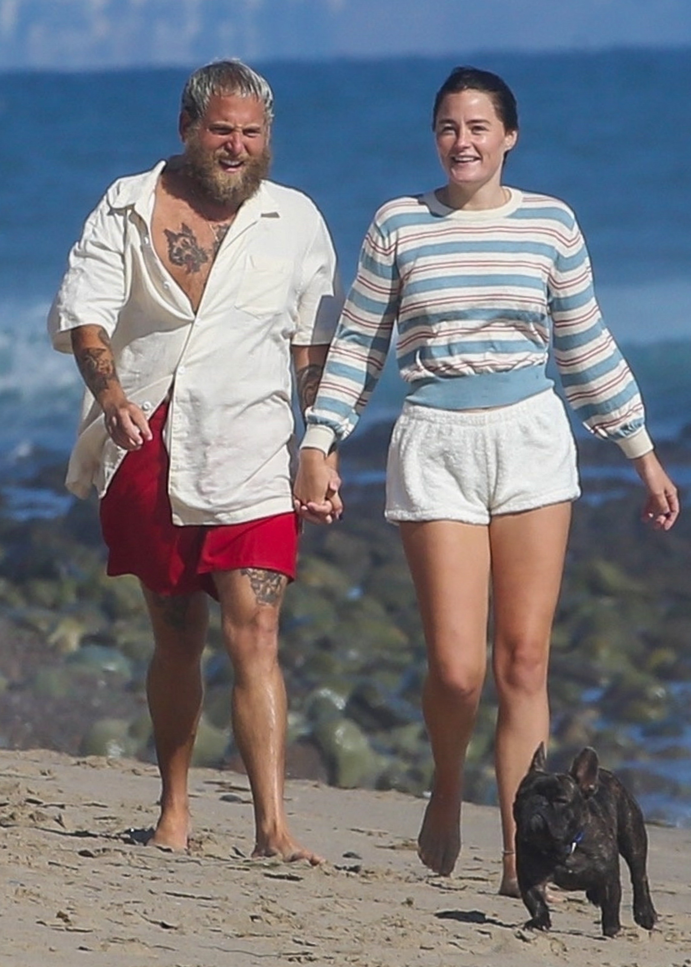 Jonah Hill and Olivia Millar holding hands while walking on the beach with a small dog.