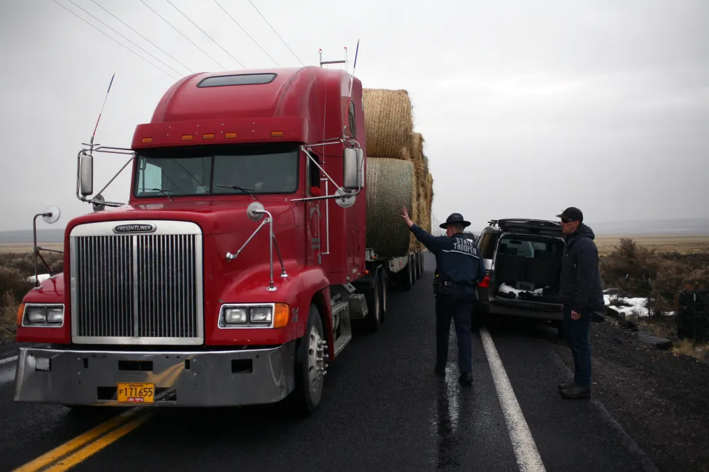 An Oregon State Trooper directing a red semi-truck loaded with hay bales past a roadblock.