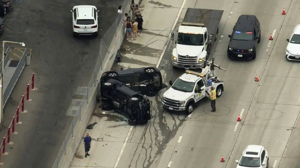 Aerial view of two black cars overturned on a highway with a tow truck nearby.