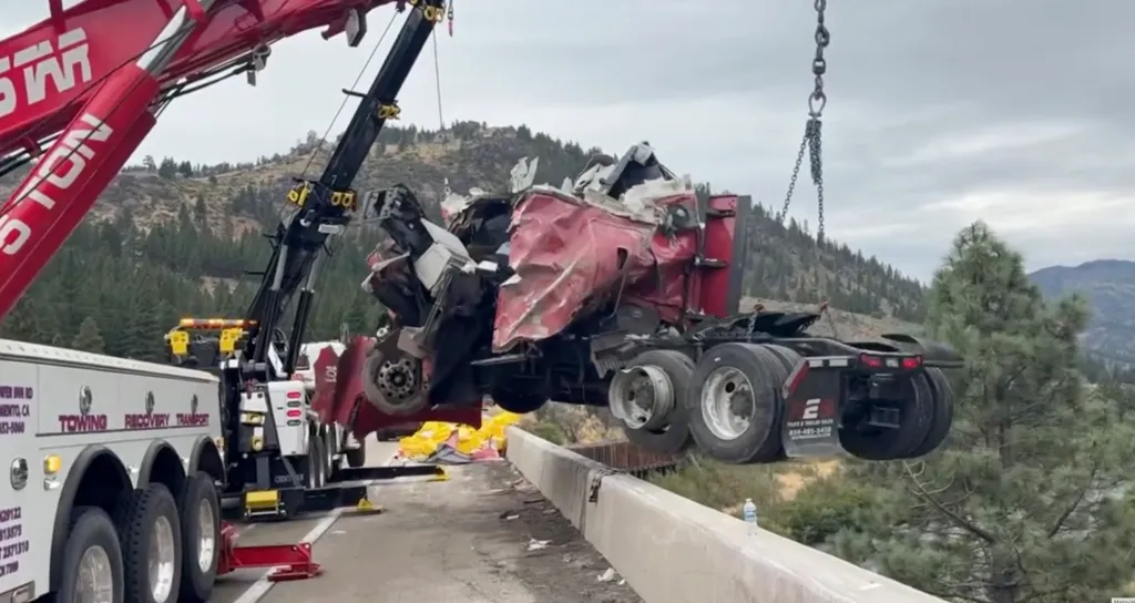 A red semi-truck being recovered by a tow truck after dangling off a bridge.