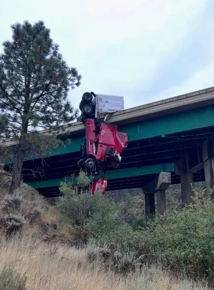 A red semi-truck crashed and is dangling upside down over the side of an interstate bridge.