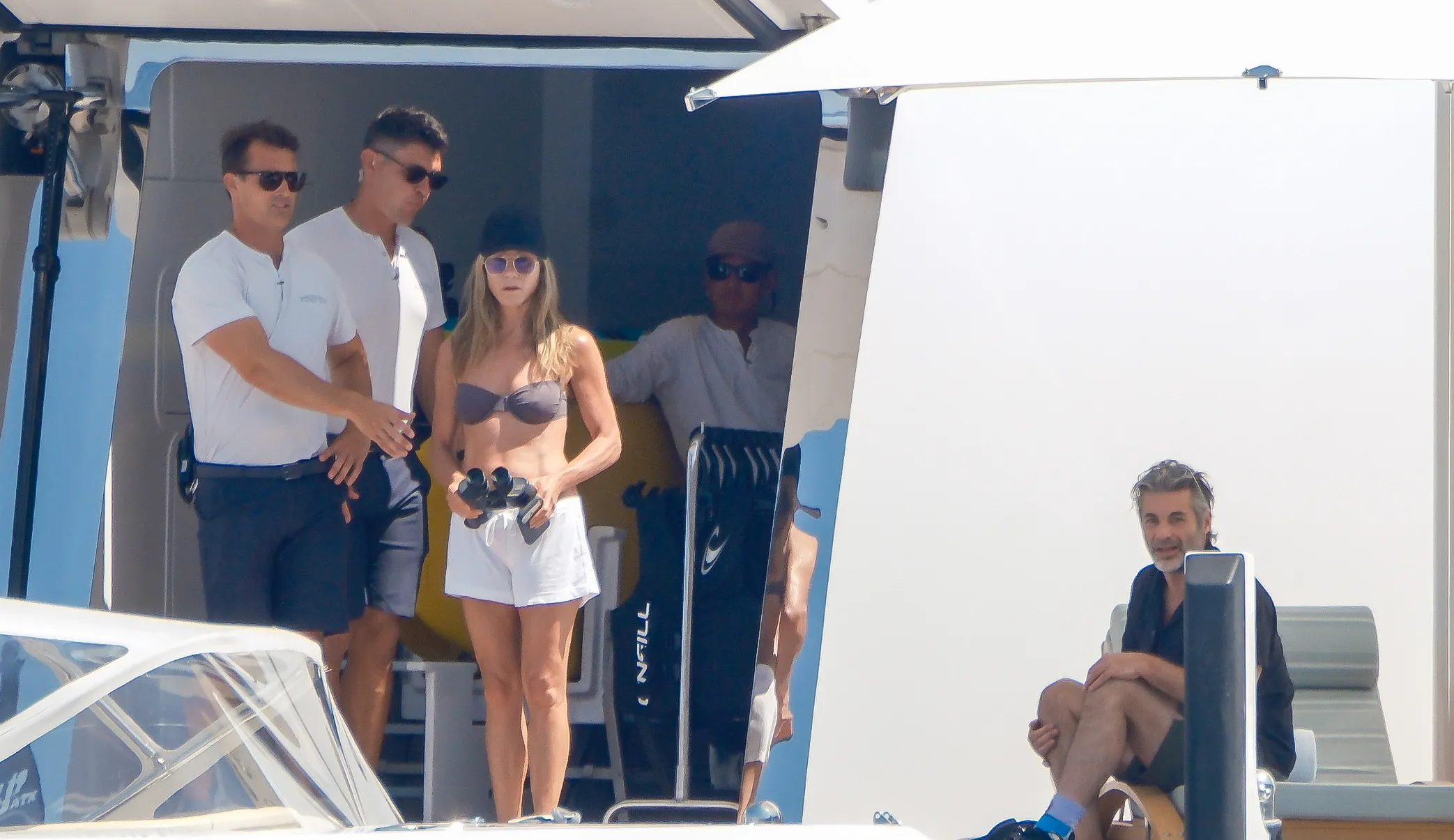 Jennifer Aniston in a bikini and shorts, holding binoculars, on a boat with friends.