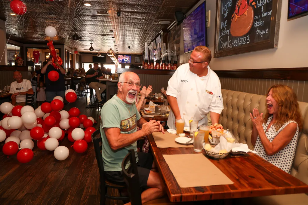 A chef presenting a lobster roll to an excited couple at a restaurant decorated with red and white balloons.