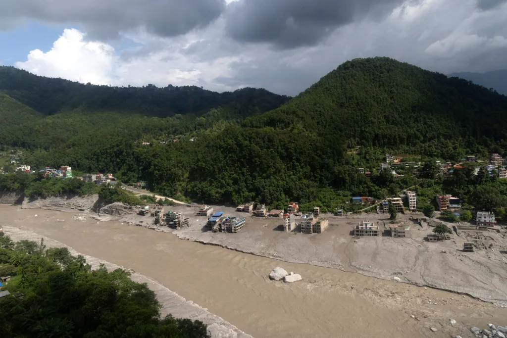 Aerial view of a Nepalese village devastated by flash floods and mudslides, with a muddy river alongside destroyed buildings and green hills in the background.