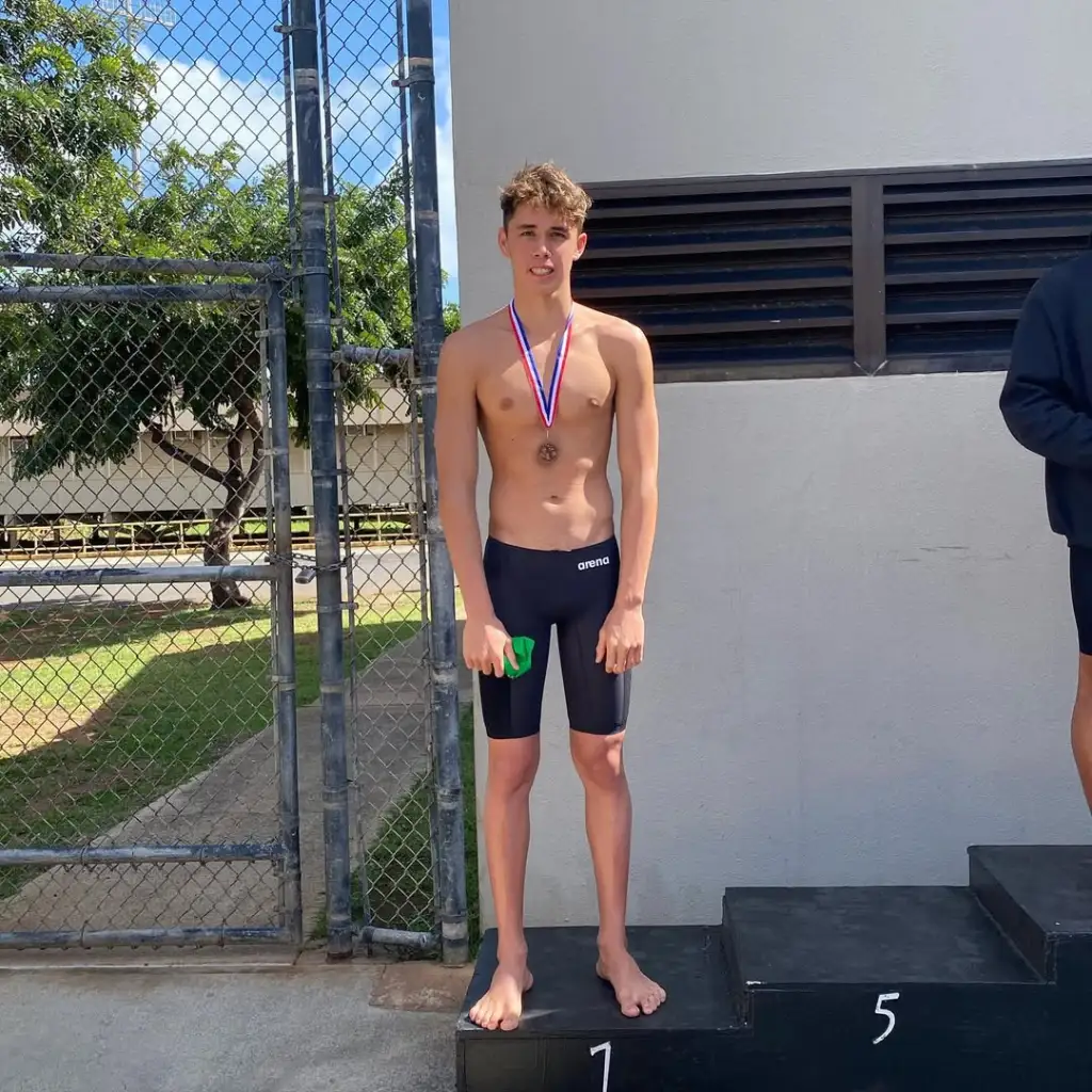A male swimmer stands on the first-place podium wearing a medal.