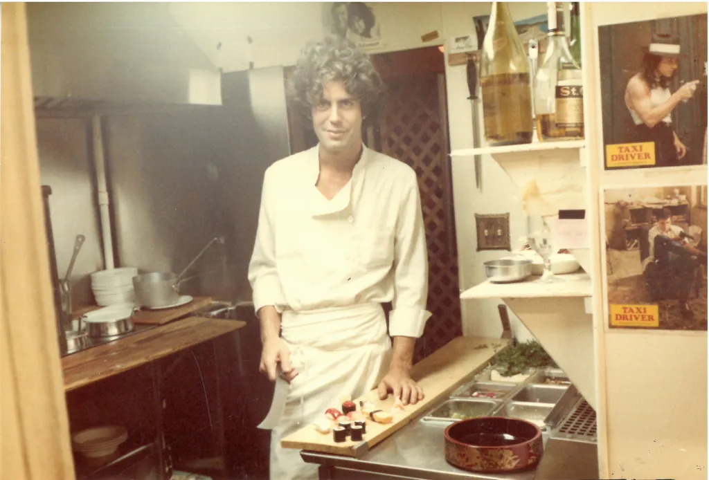 Tony Bourdain in a kitchen, wearing a chef's jacket and apron, preparing sushi.