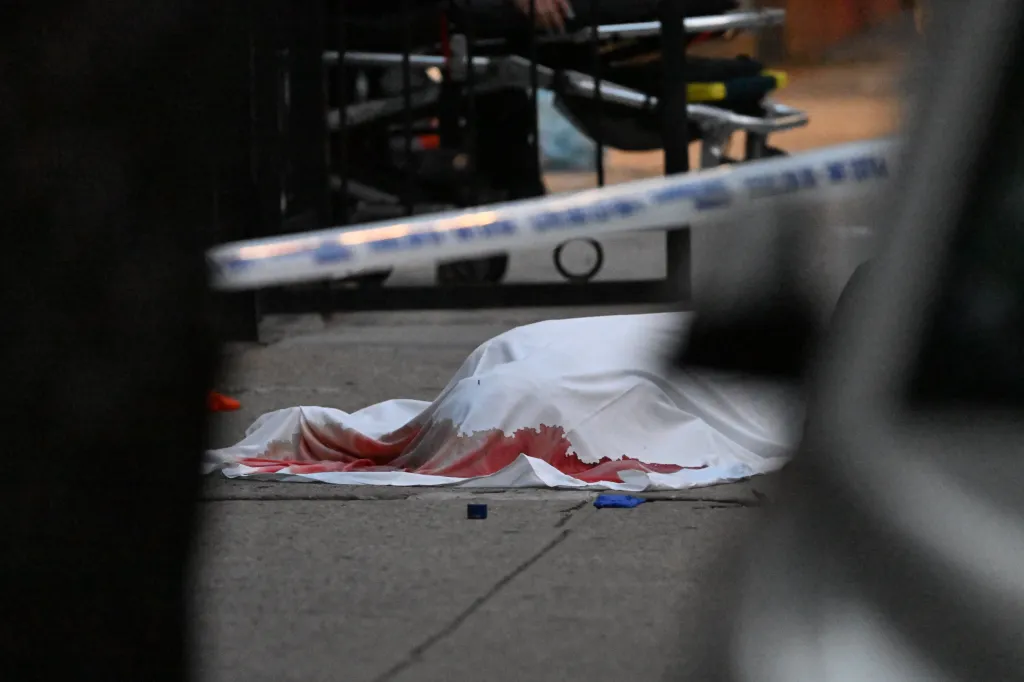 A white sheet covers a body on the ground, stained with blood, with crime scene tape visible in the foreground.