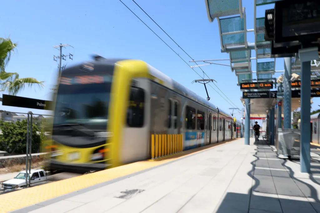 A light rail train speeding past a station platform under a bright blue sky.