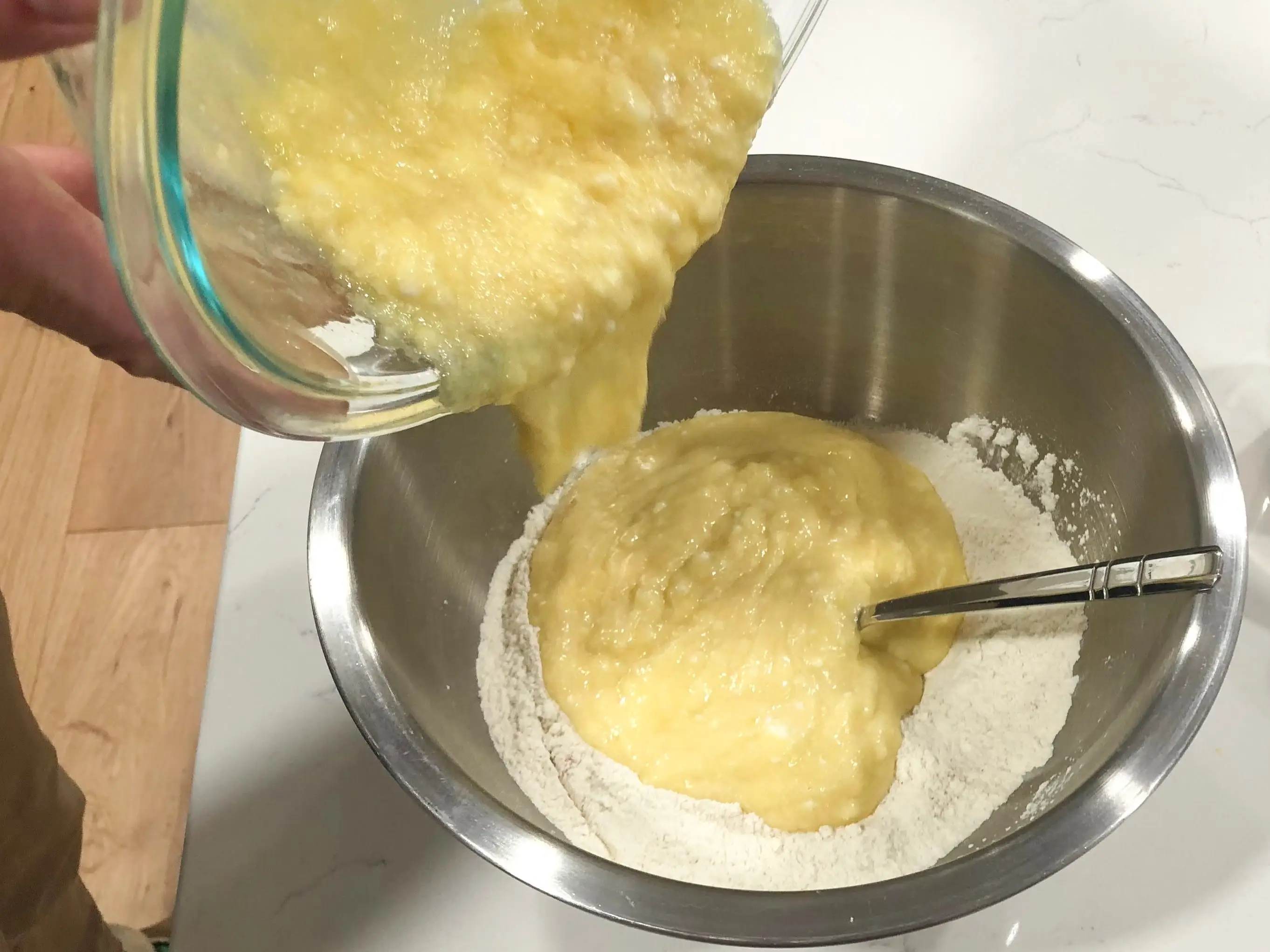 A person pours the wet ingredients from the clear bowl into the dry ingredients in the stainless-steel bowl. There are also measuring cups and a spoon on the counter.