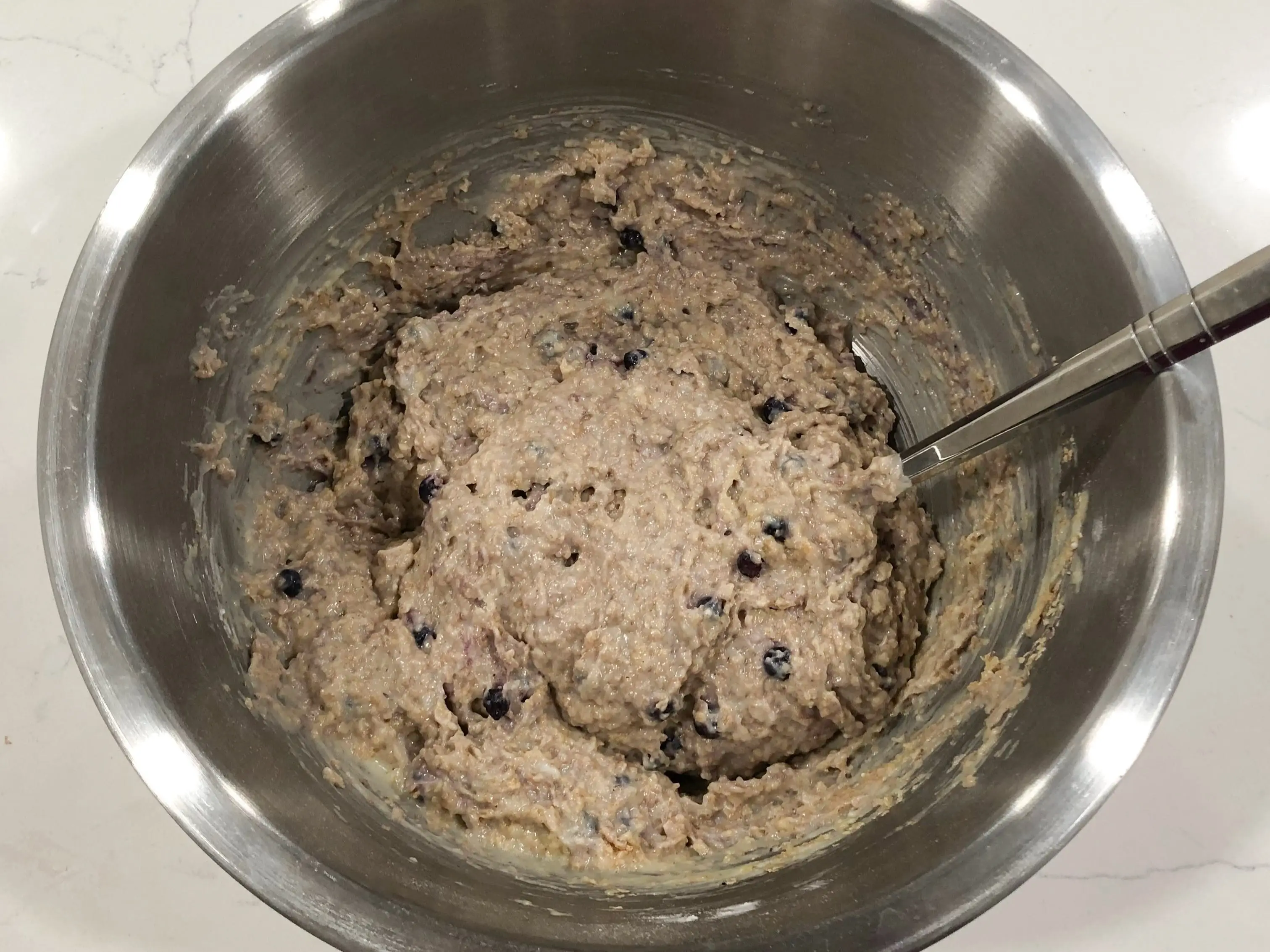 Muffin batter, which now includes blueberries and is a darker color, in a stainless-steel bowl with a spoon.