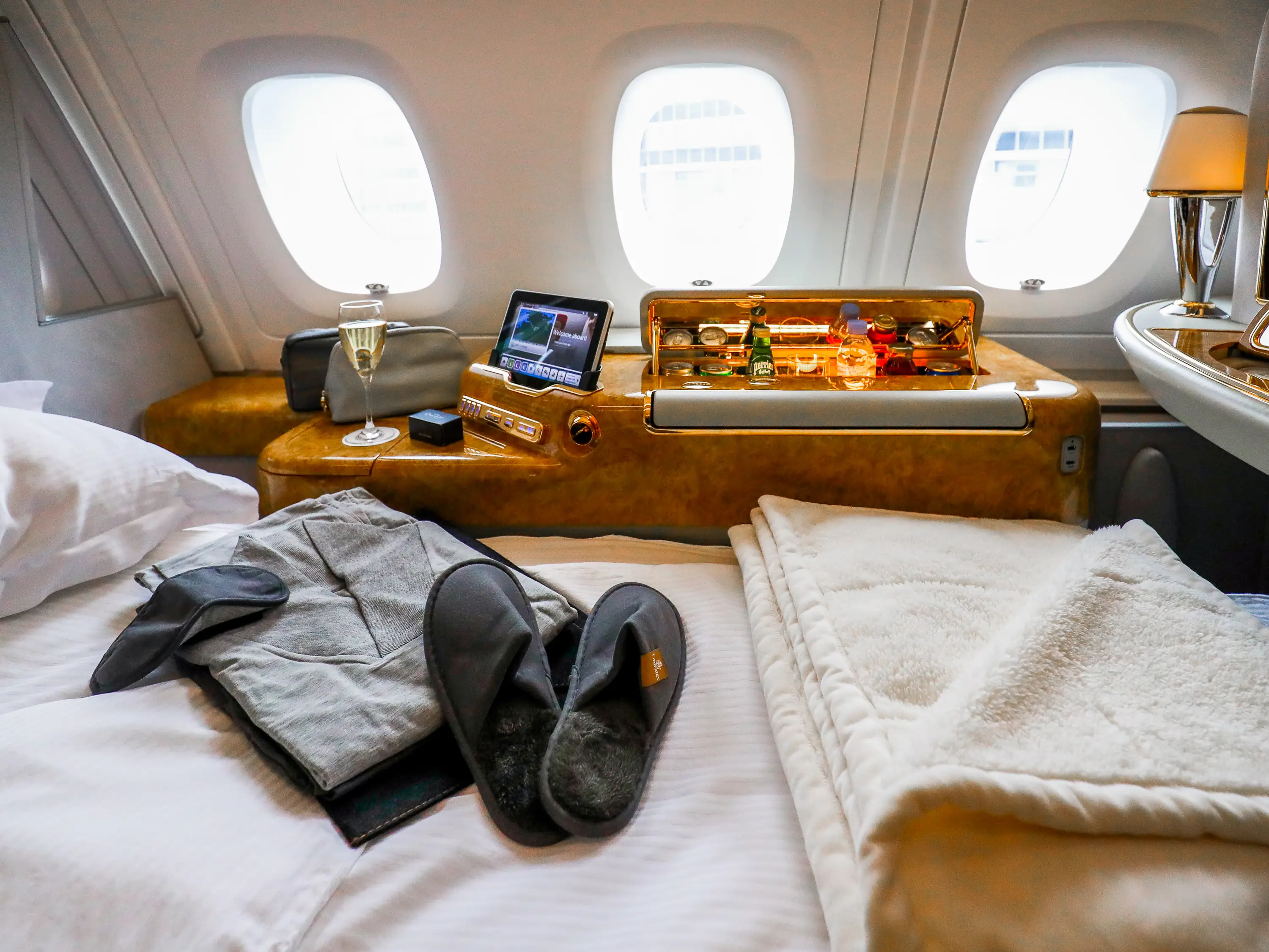 A bed, slippers, a blanket, a robe, and a sleep mask on a bed inside the first class cabin inside an Airbus A380 at the airbus factories in Hamburg, Germany, Refreshments in the background in front of three windows