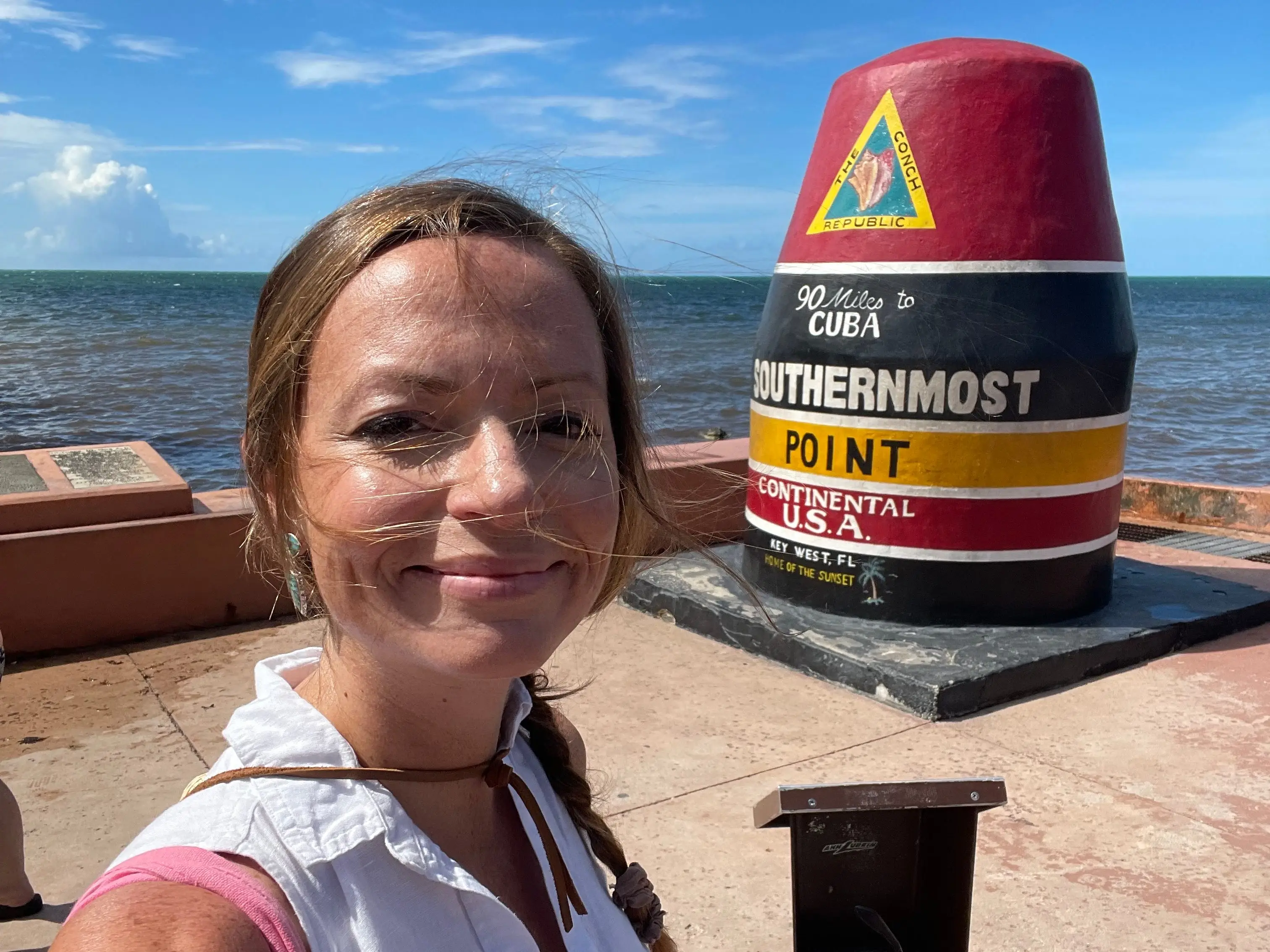 Emily taking a selfie in front of the Southernmost Point in Key West, Florida, with the ocean in the background.
