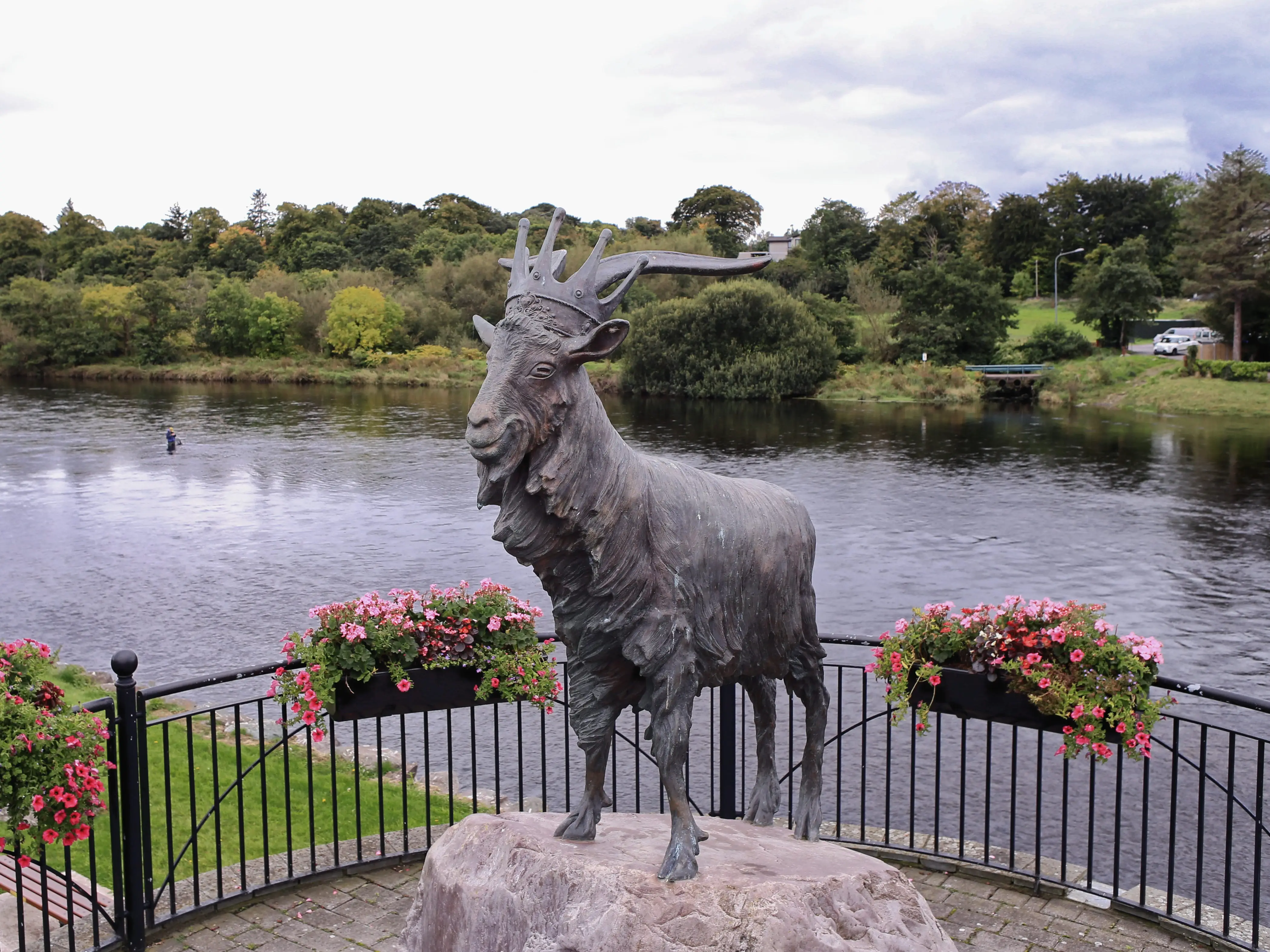 A statue of a goat wearing a crown surrounded by a black fence with pink flowers in boxes in front of a body of water.