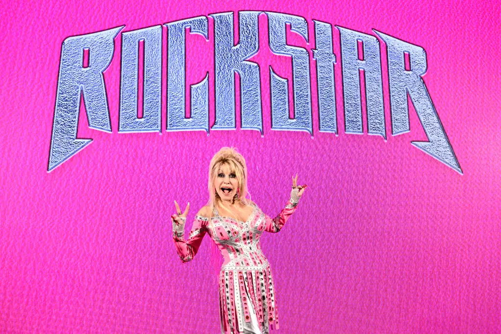 Dolly Parton poses in front of a screen that reads 