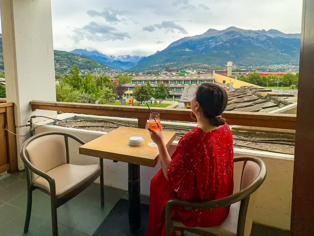 The author sitting with her back faced to the camera at a table on a balcony in Valle d'Aosta, Italy; she's holding a drink in her left hand as she looks out at the view.