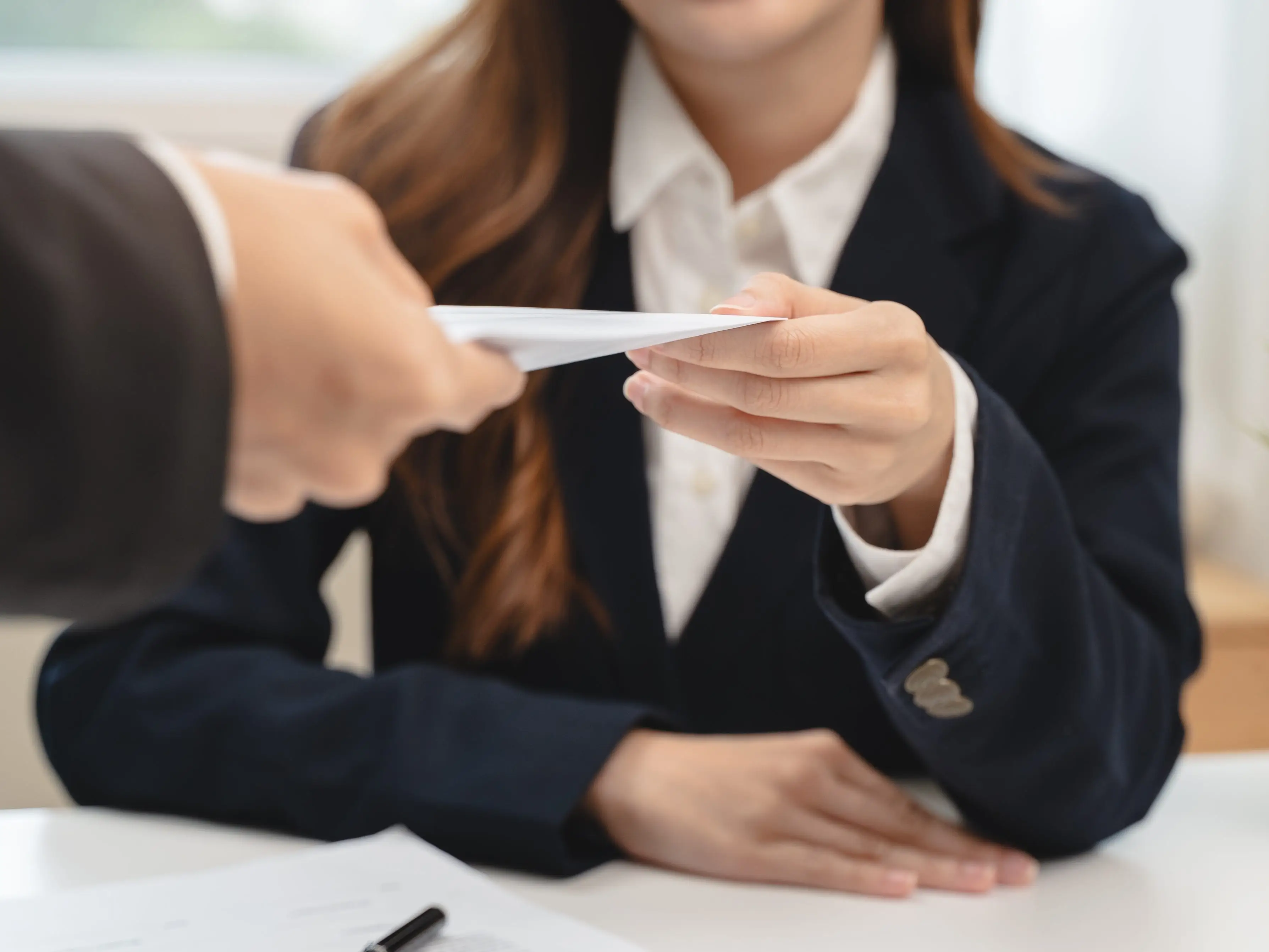A person hands an envelope across a table to a woman wearing a suit.
