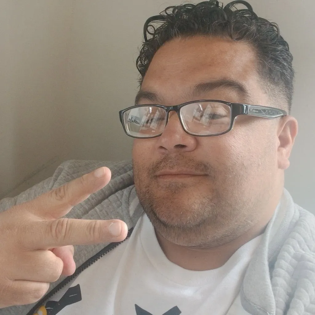 A man wearing glasses and a gray hoodie flashes a peace sign.