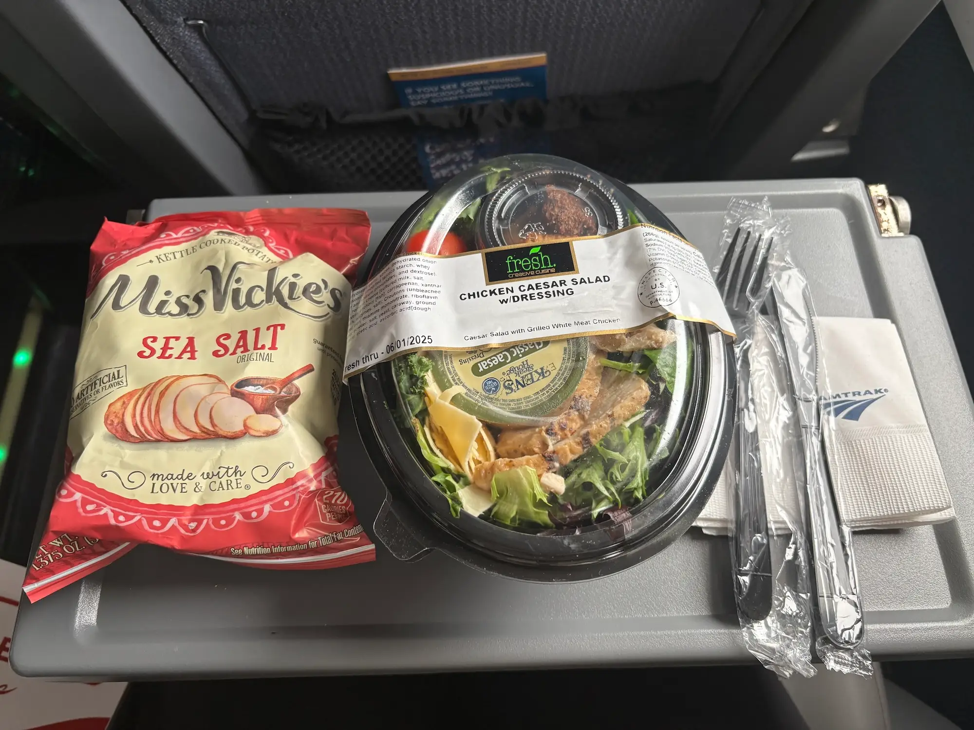 A bag of chips, a pre-packaged caeser salad, plastic silverware, and an Amtrak napkin on a tray table on a train.