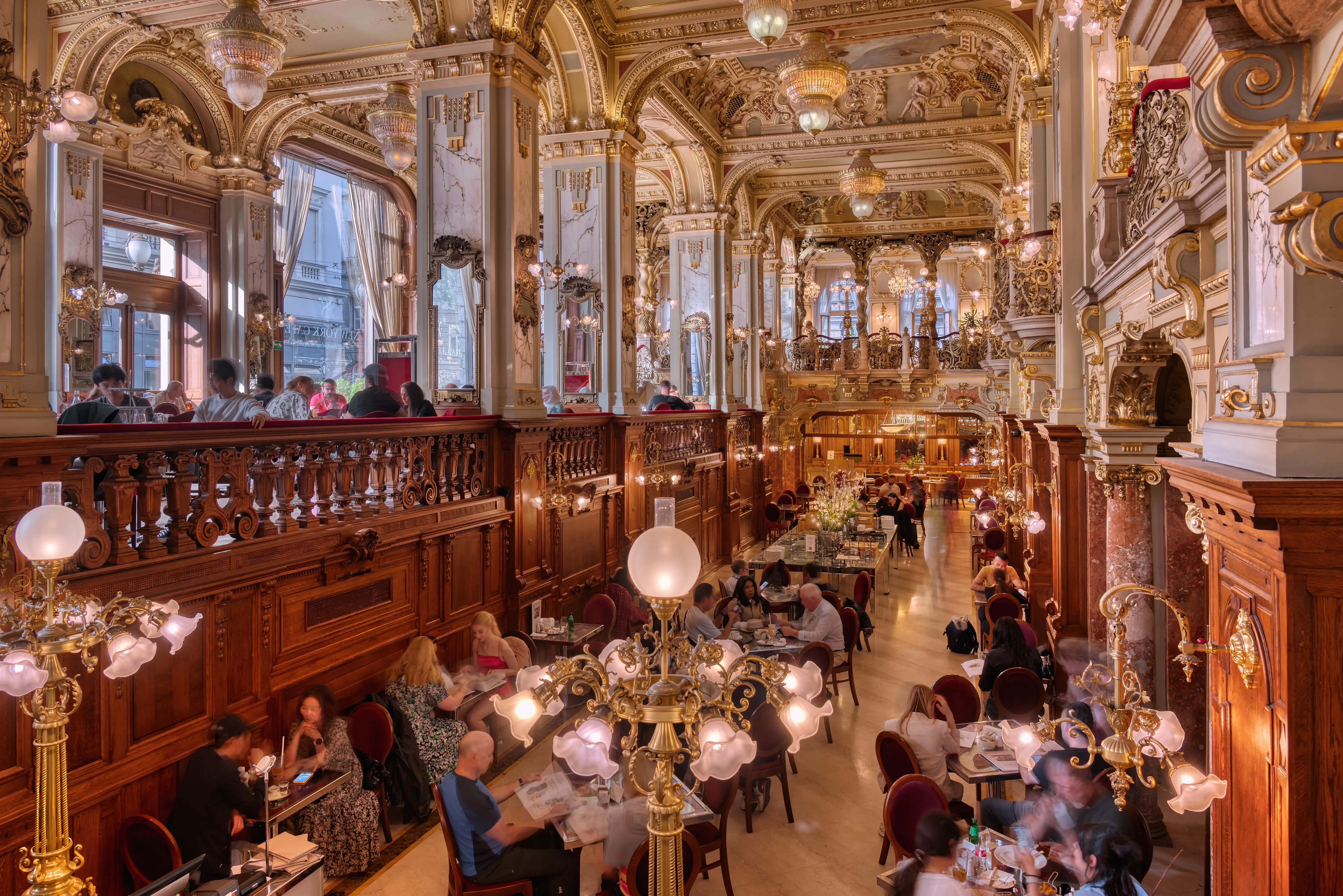 The interior of the New York Cafe in Budapest, an ornately decorated coffeehouse with people sitting at round tables.