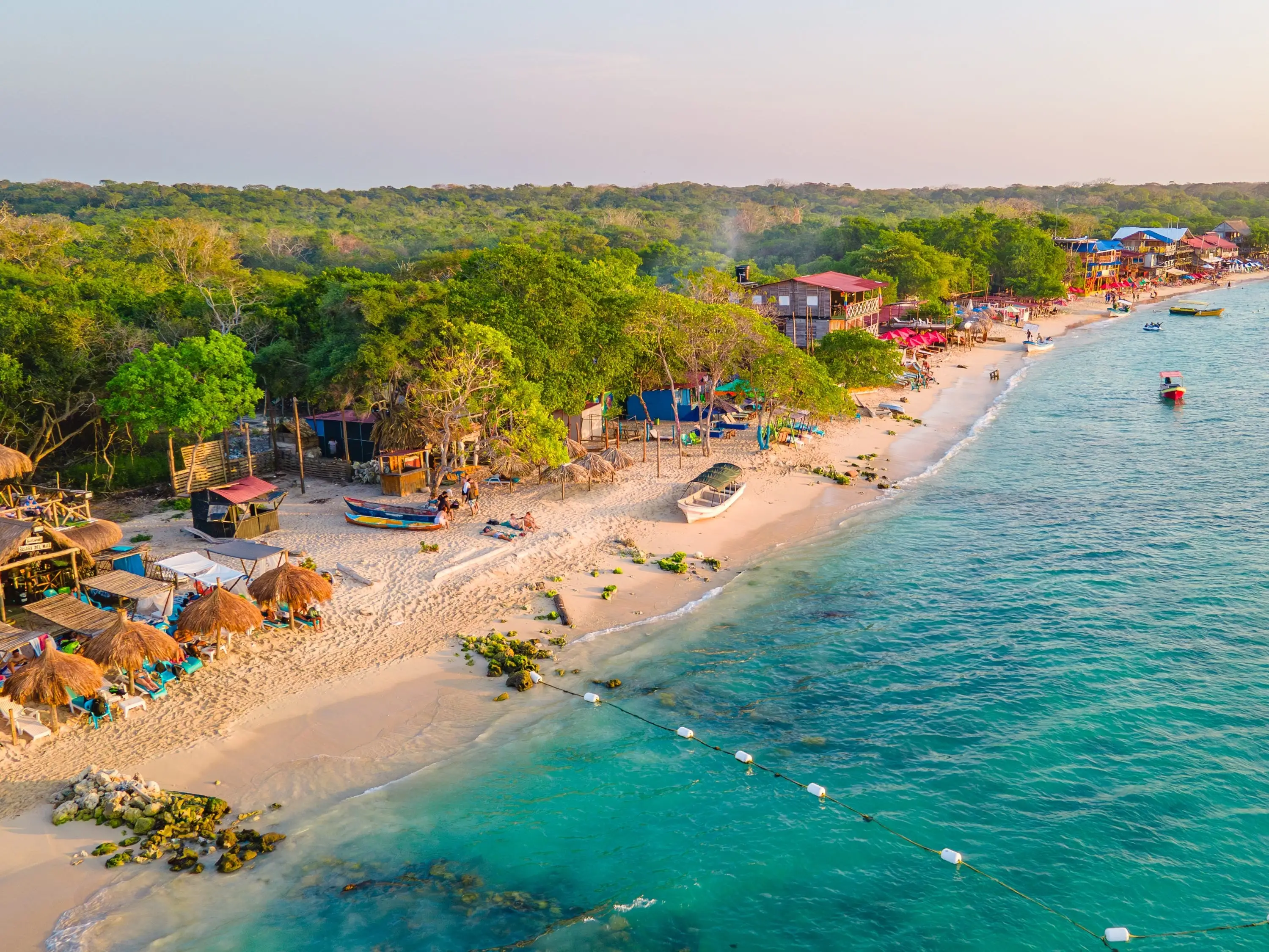 Aerial view of Playa Blanca, Baru with clear blue waters, sandy beach, boats, and rustic buildings amidst green trees in Cartagena, Colombia