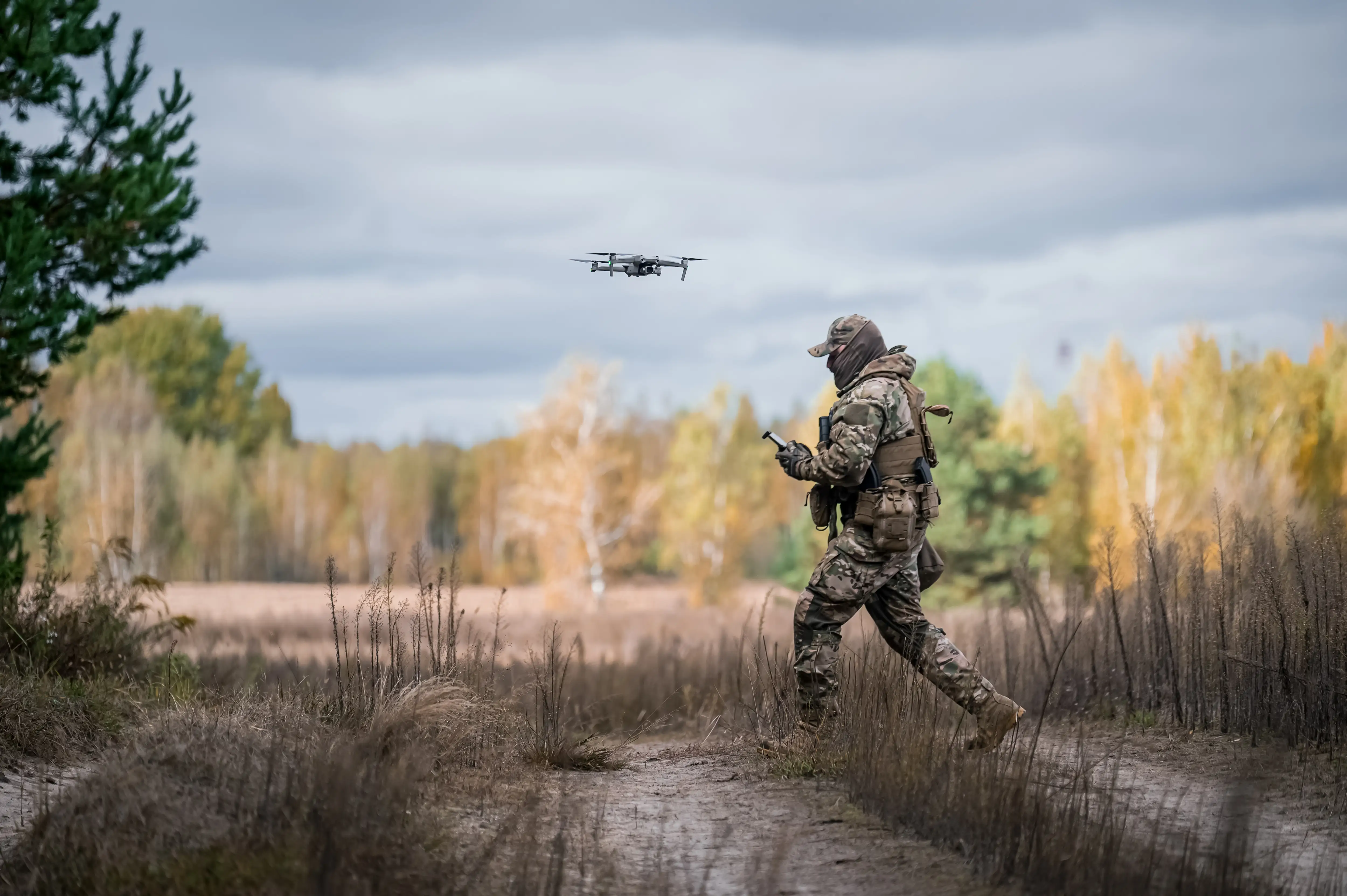 A man in camouflage gear walks in a field with a small drone in front of him and trees behind him