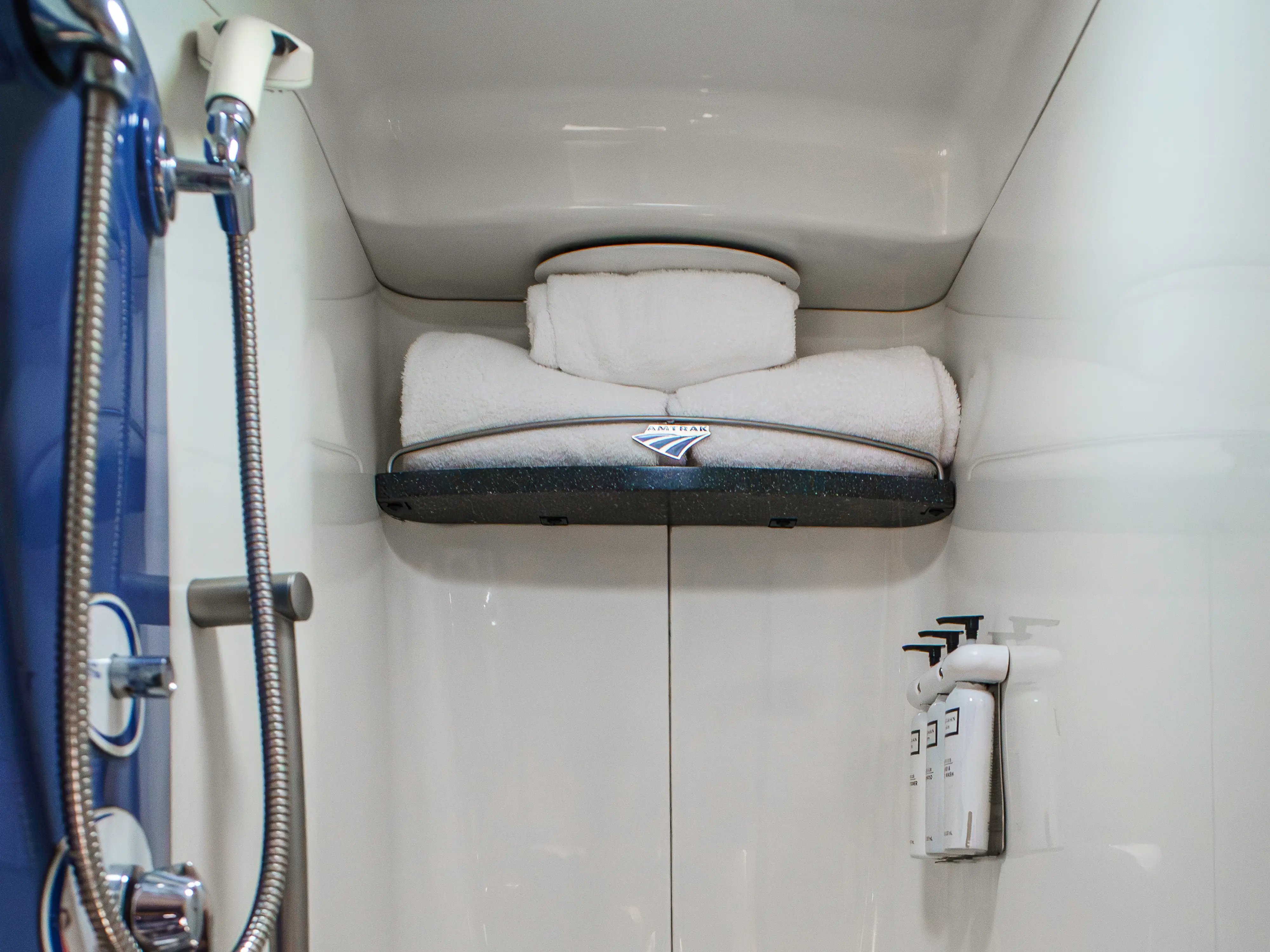 Three towels on a shelf in an Amtrak shower with the shower head on the left and toiletries on the wall on the right