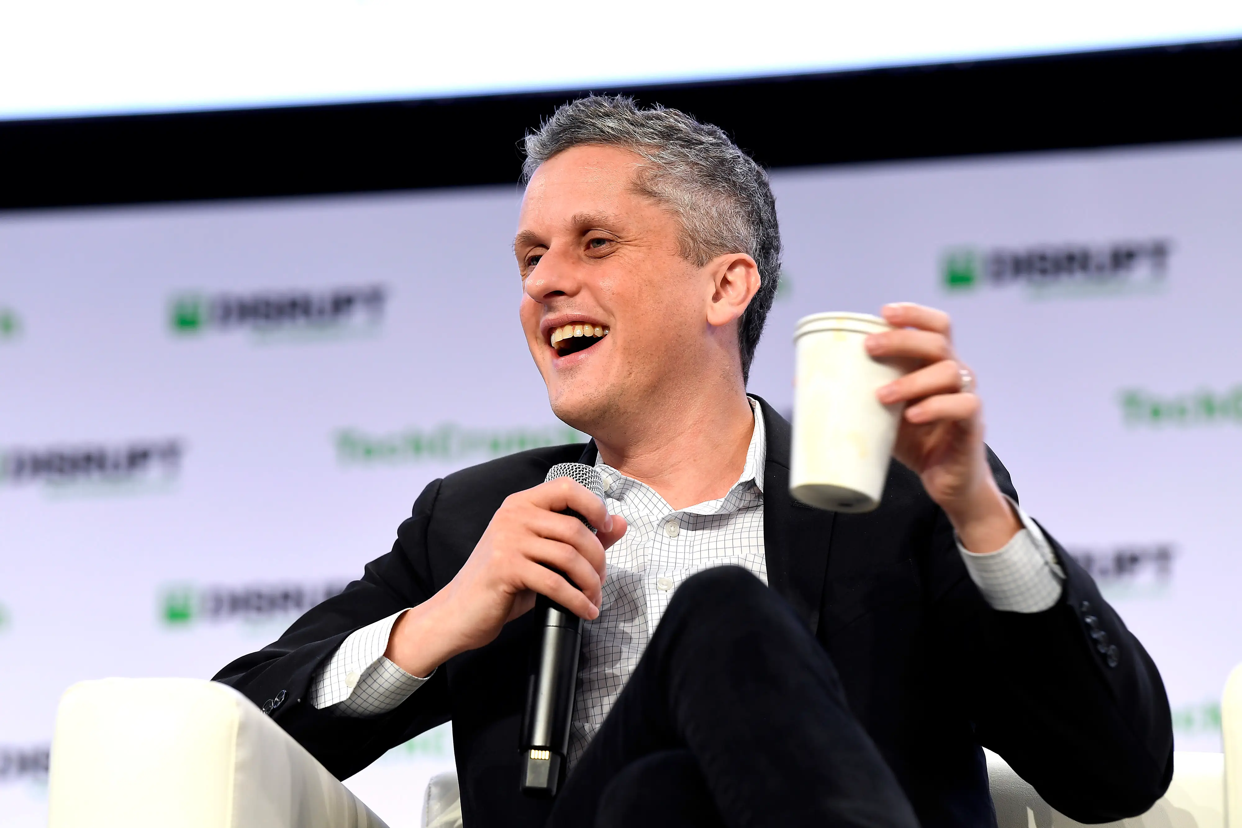 Aaron Levie, dressed in a dark suit jacket and a light button-up, holds a mic and a coffee cup onstage.