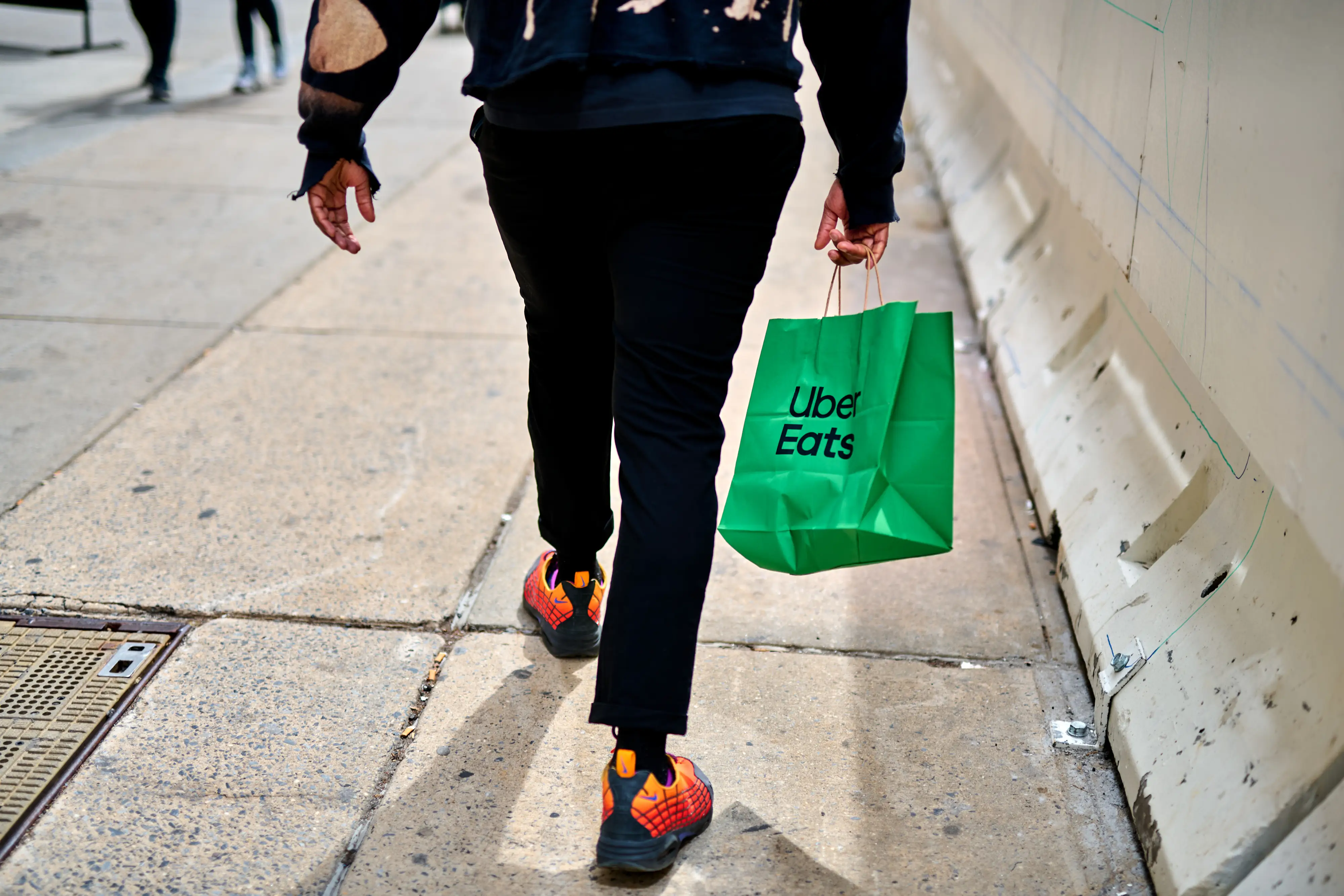 A person walks away from the camera carrying a green paper Uber Eats bag while wearing dark pants and red, yellow, and black sneakers.