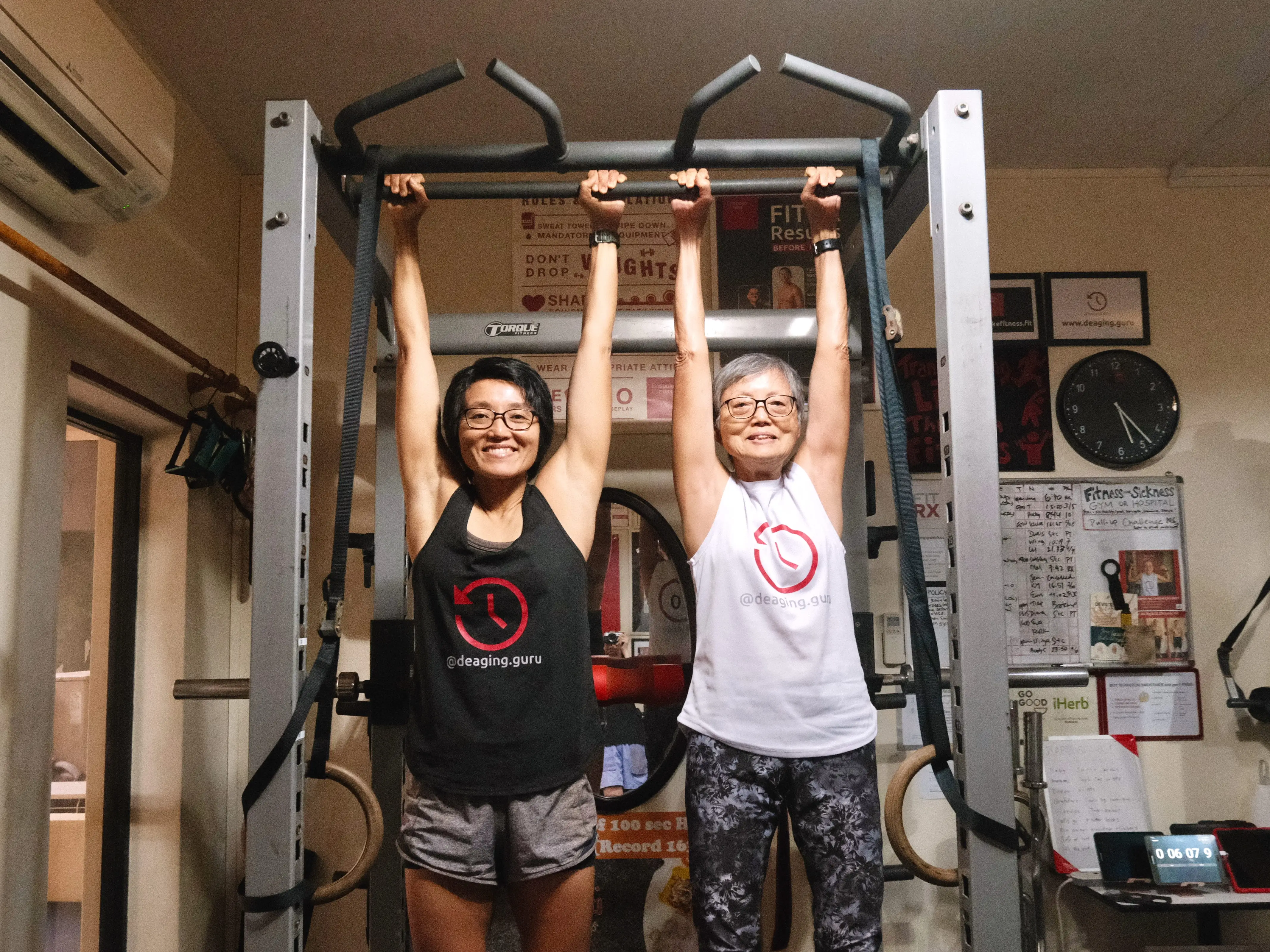Two women are doing a dead hang on a pull-up bar.