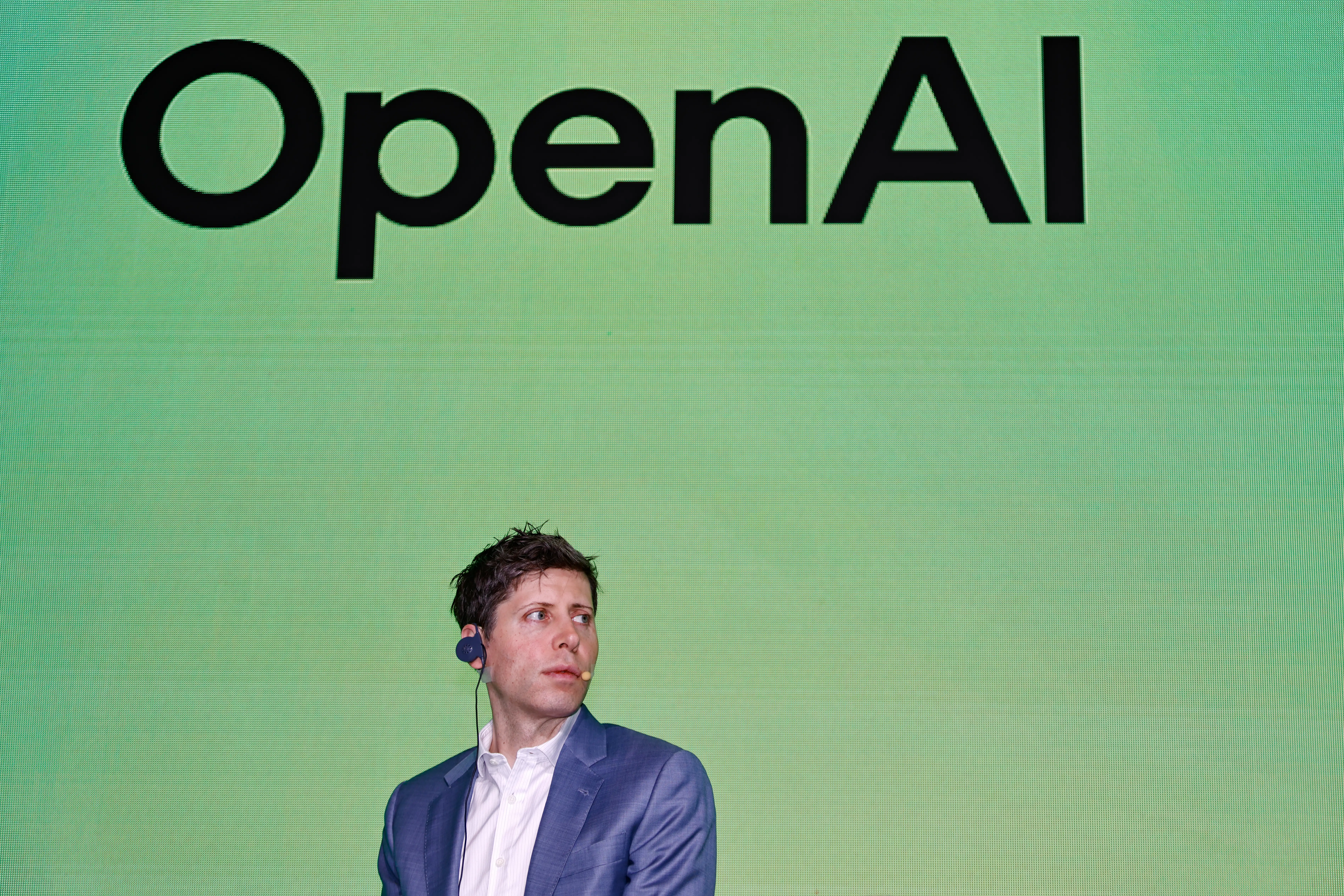 Sam Altman sits below a large OpenAI wordmark on a green presentation screen.
