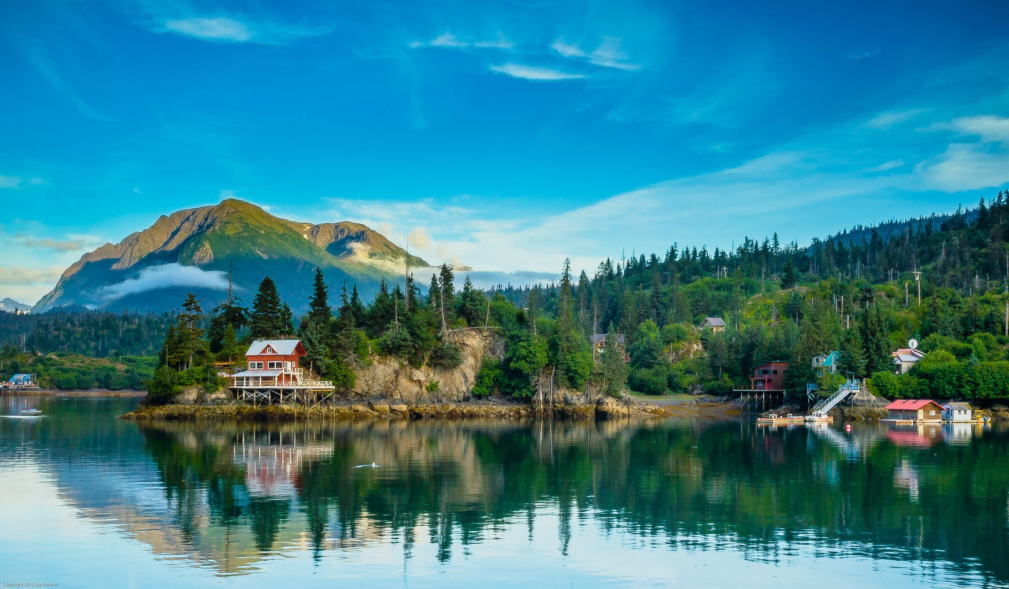 Coastal houses sit along a forested shoreline with mountain peaks and reflections on calm blue water.