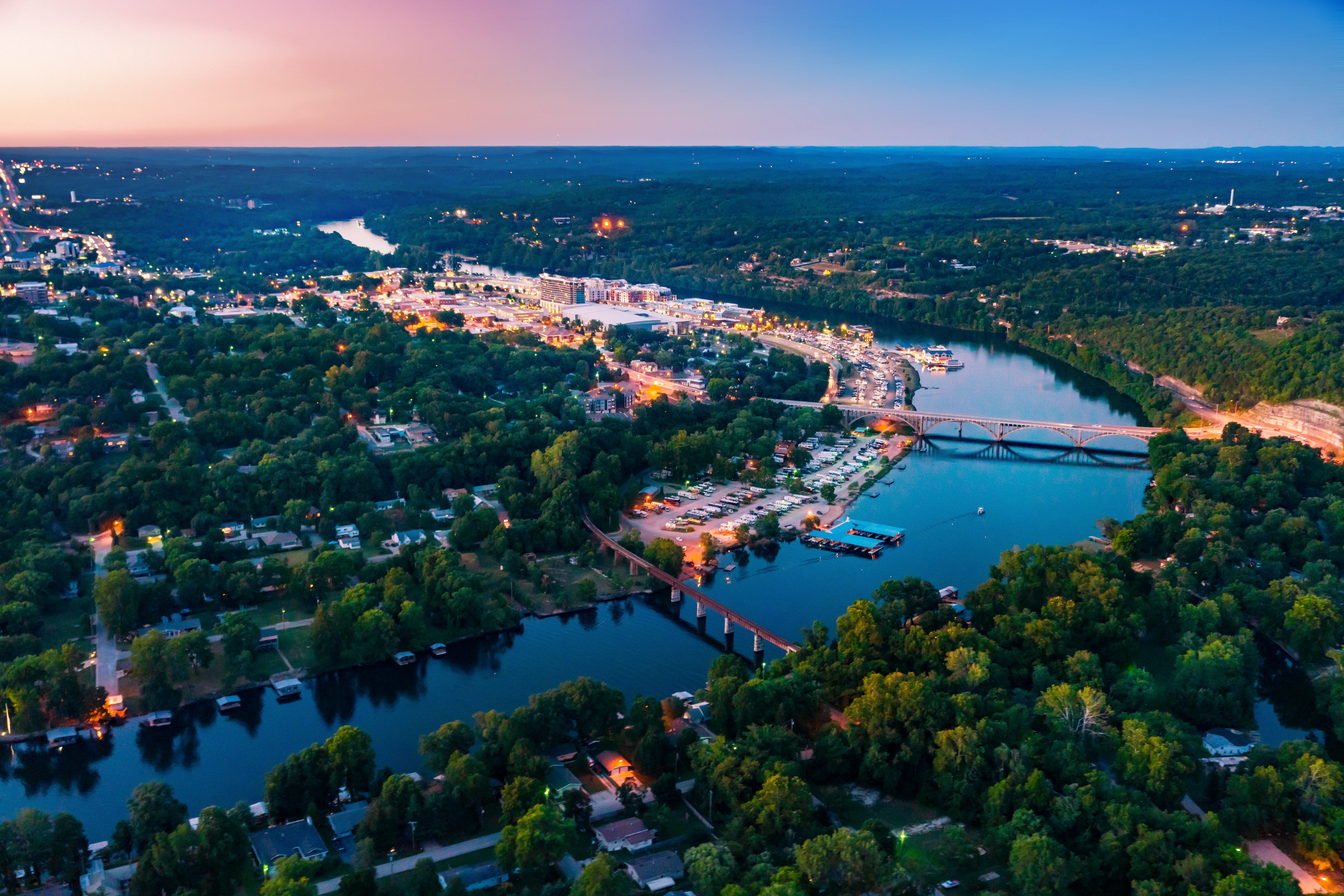 Aerial twilight view of a riverfront town with bridges, marinas, wooded neighborhoods, and city lights.