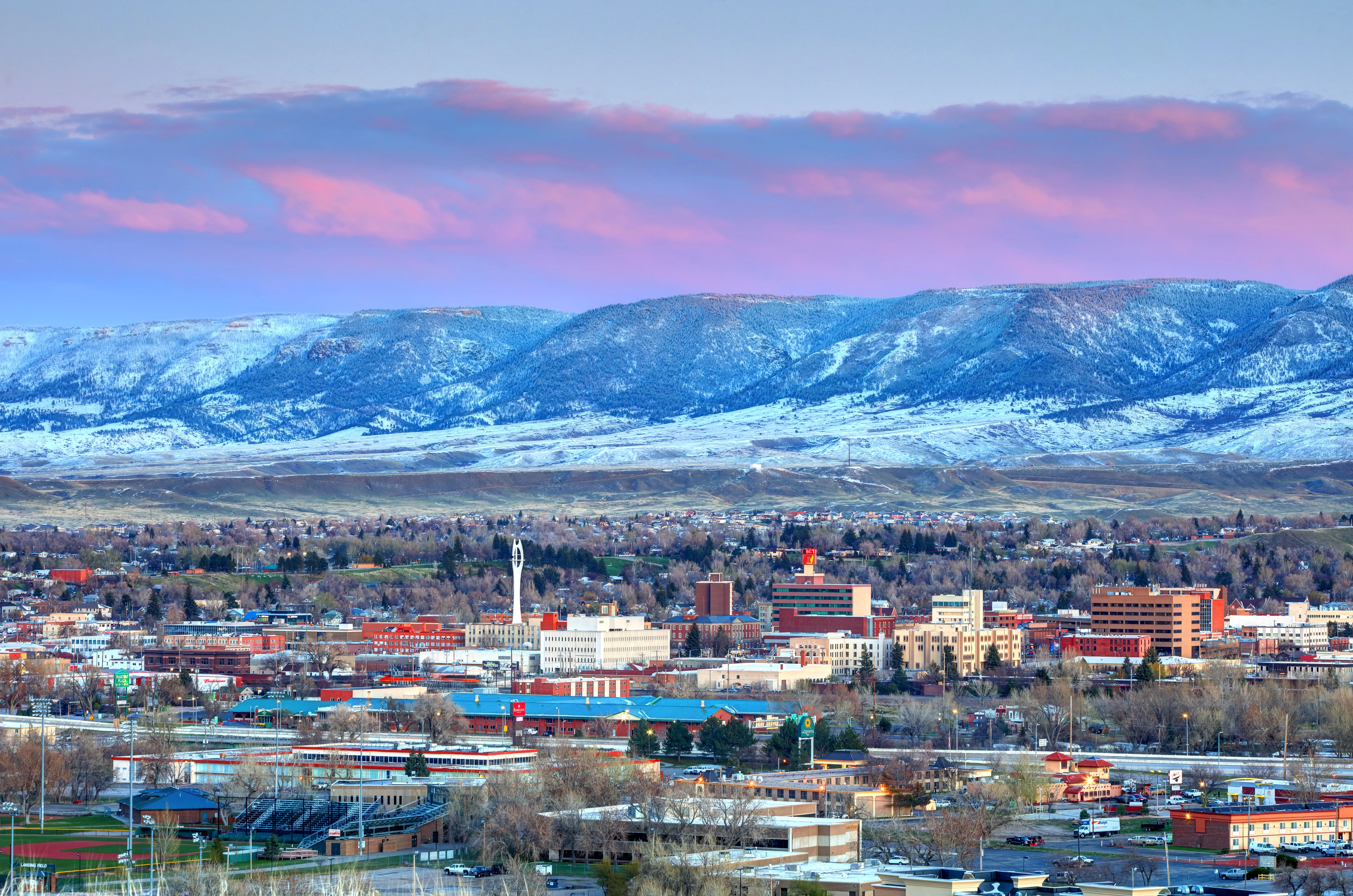 Snow-dusted mountains rise behind a city skyline under pink clouds at sunset.
