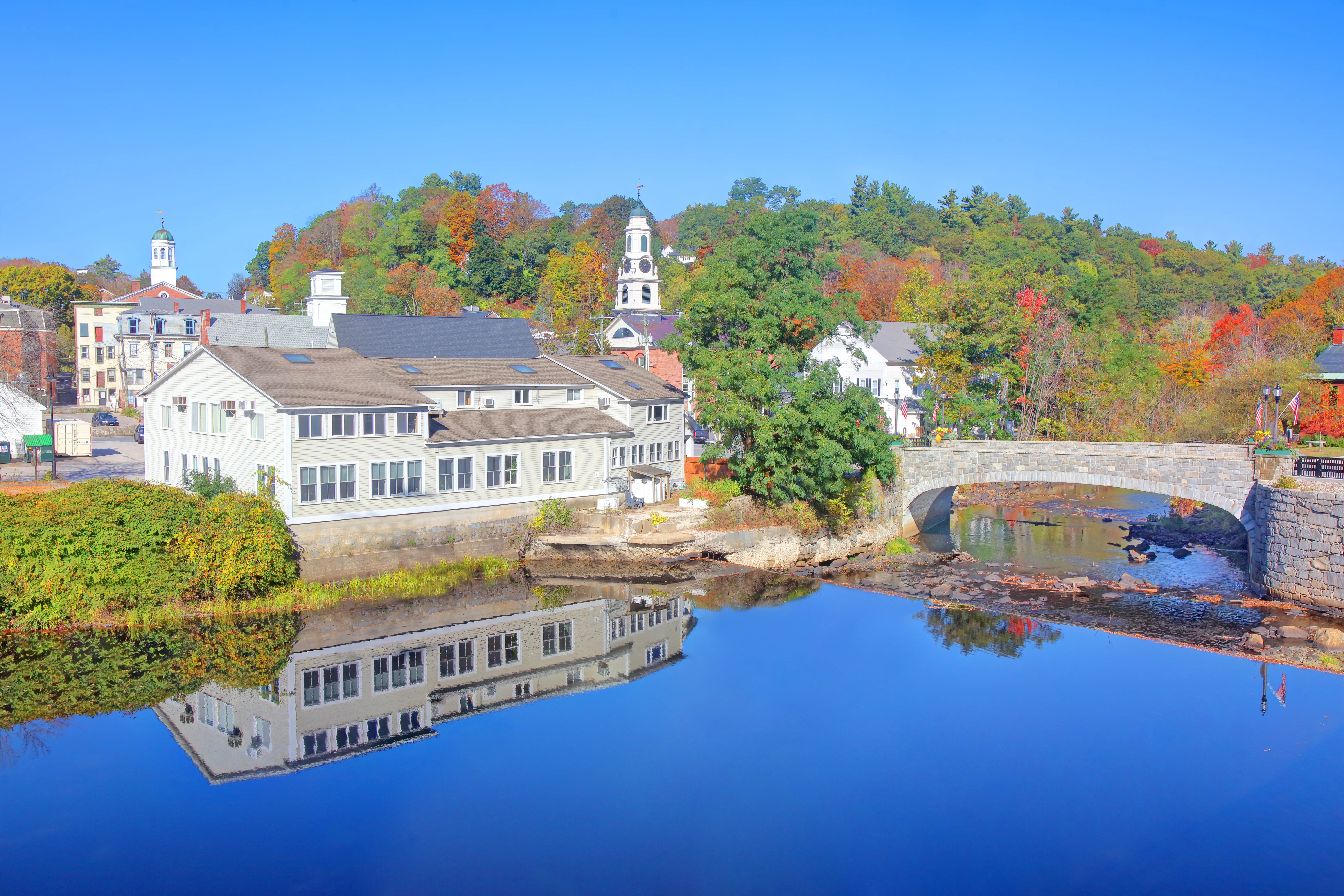 Autumn trees, white riverside buildings, church steeples, and a stone bridge reflect in calm blue water.
