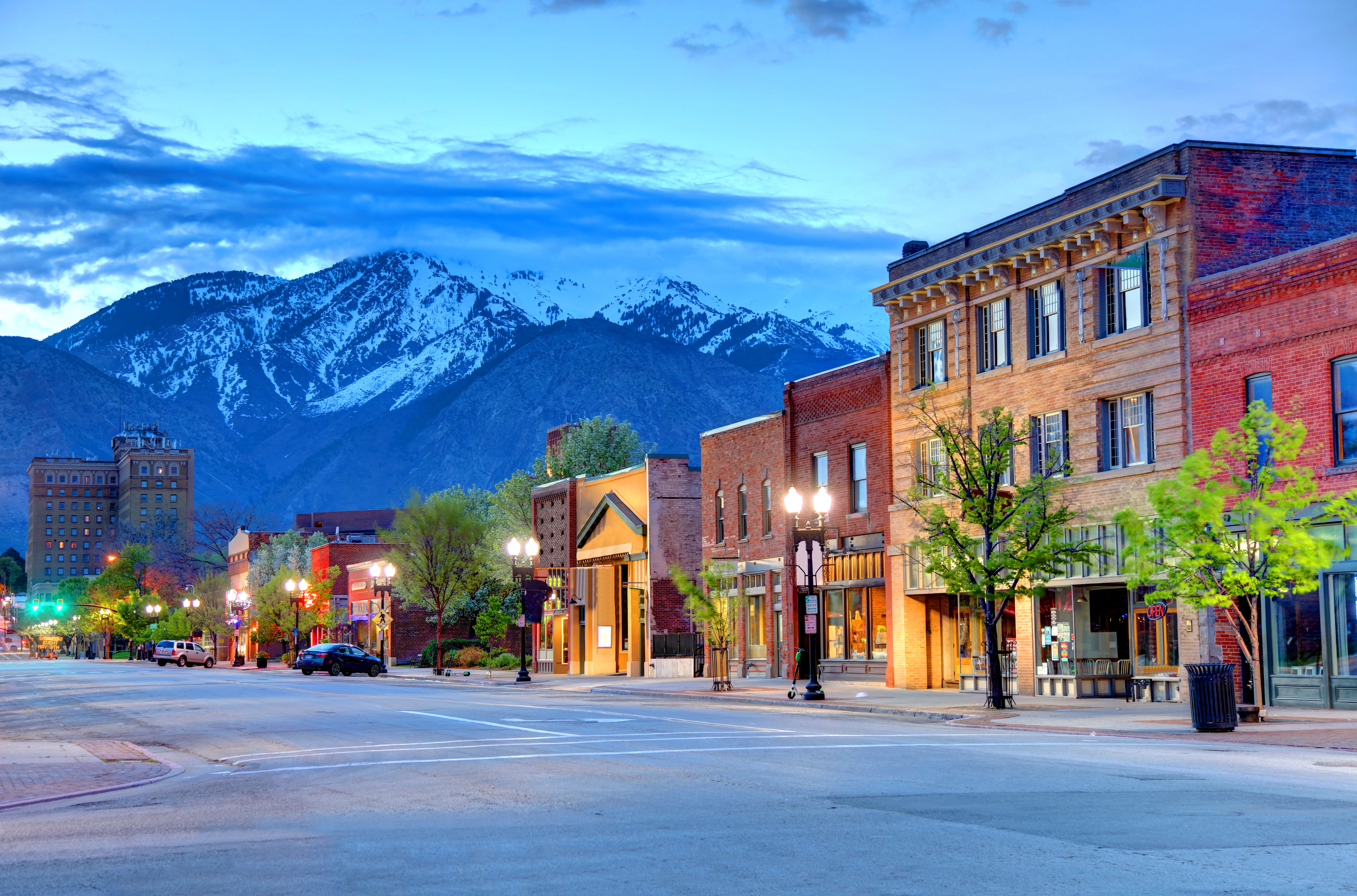 Downtown street with lit storefronts at dusk beneath snow-capped mountains and a blue evening sky.