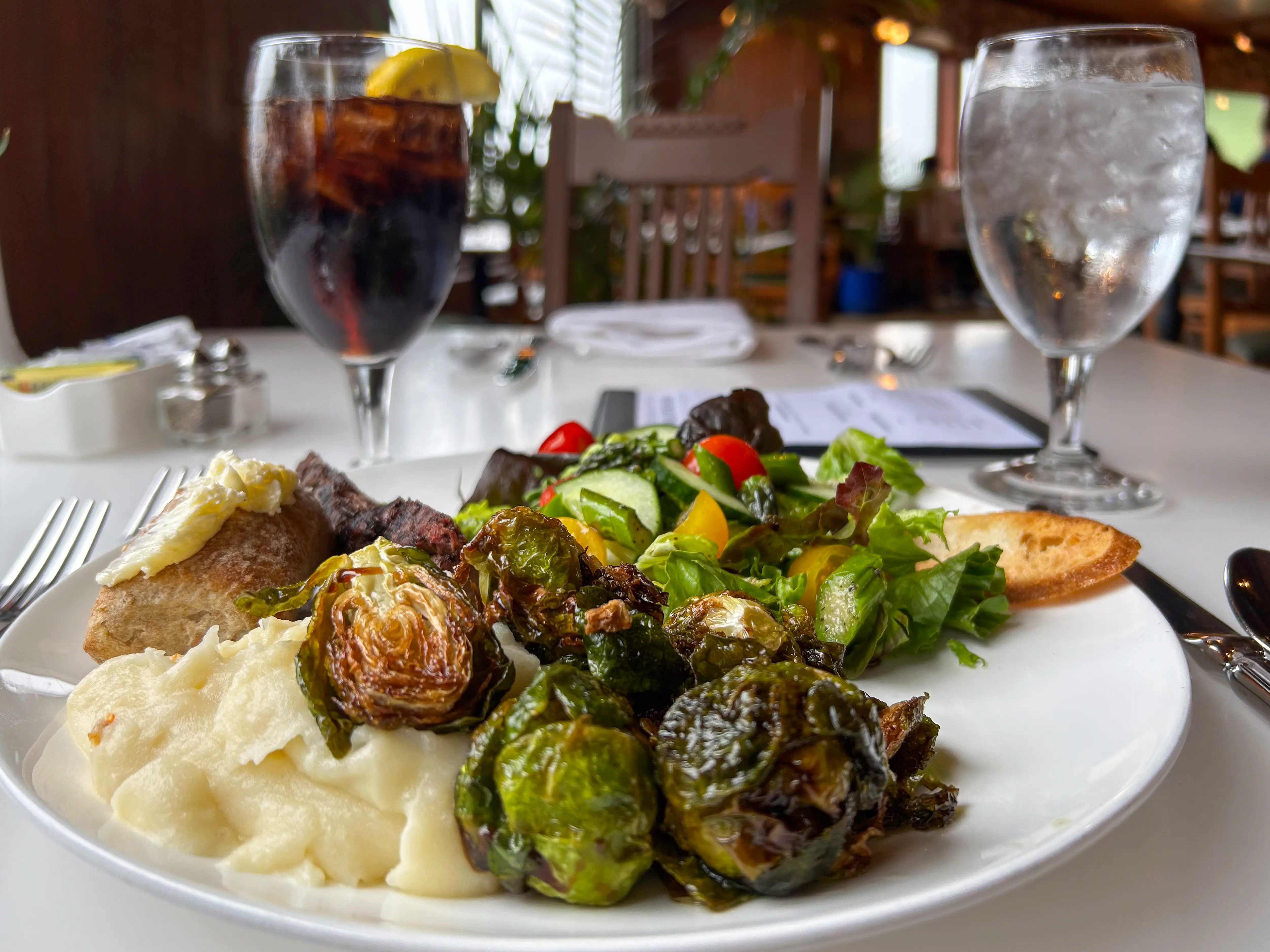 Restaurant table setting with plated steak, mashed potatoes, roasted Brussels sprouts, salad, toast, and two drinks.