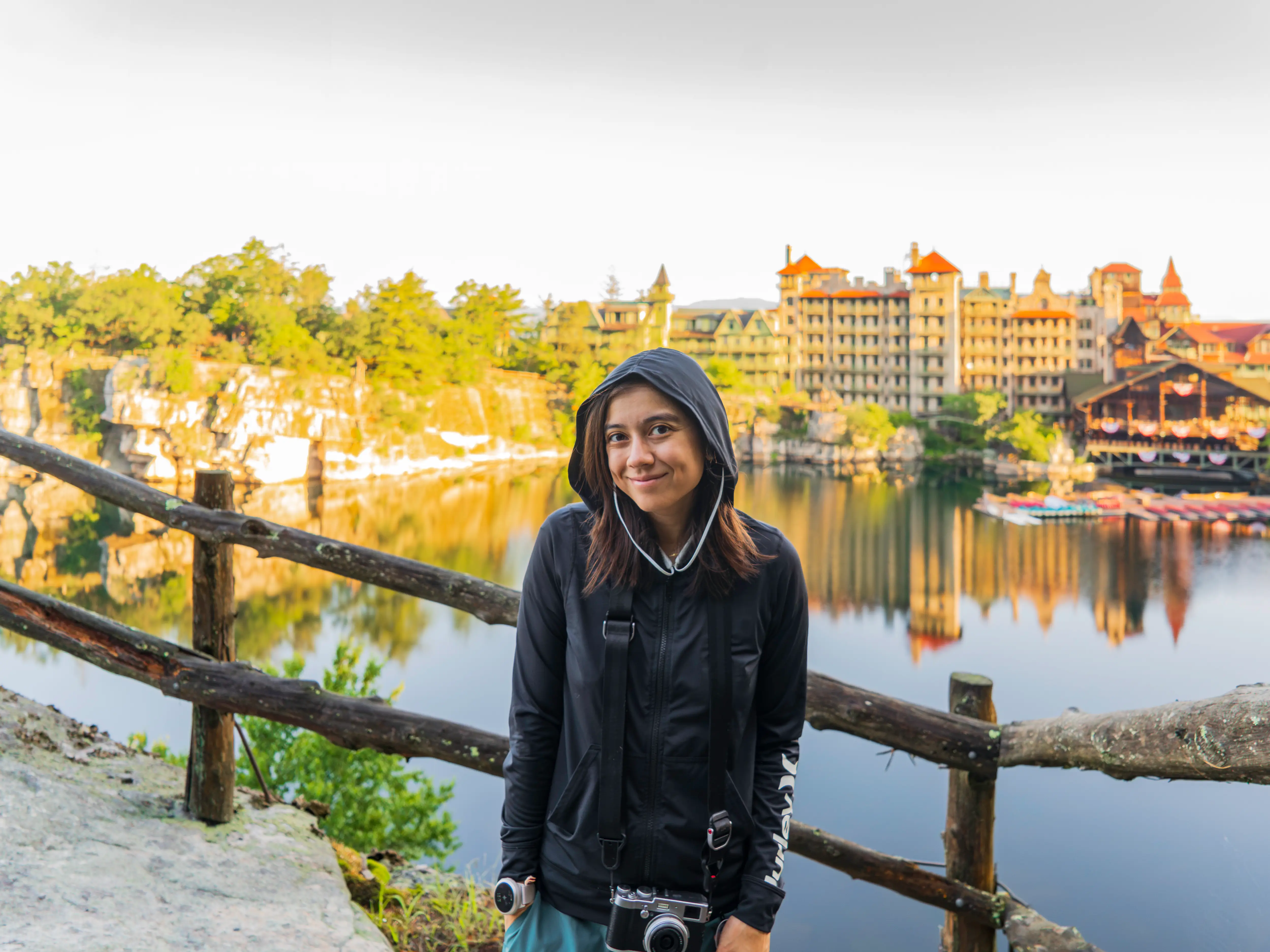 Hooded visitor with a camera stands by a wooden fence overlooking a lake and castle-like resort, Mohonk Mountain House