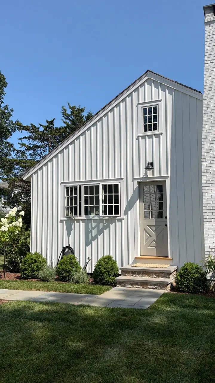 A white cottage with a taupe door in a yard with greenery.